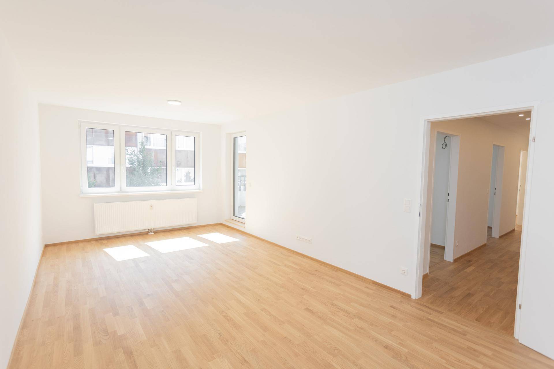 Helles, unmöbliertes Zimmer mit Holzfußboden und Zugang zu einem Flur, der weitere Räume erschließt.