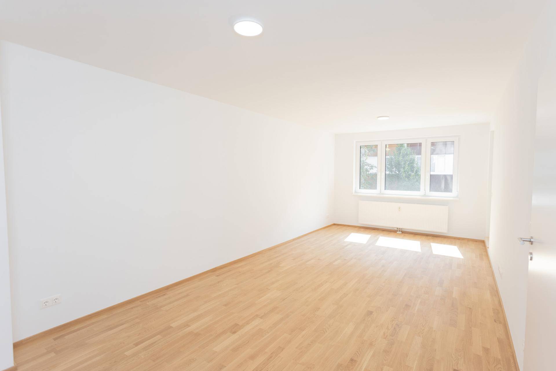 Helles, leeres Zimmer mit weiß gestrichenen Wänden und einem warmen Holzfußboden, durchflutet von Sonnenlicht.