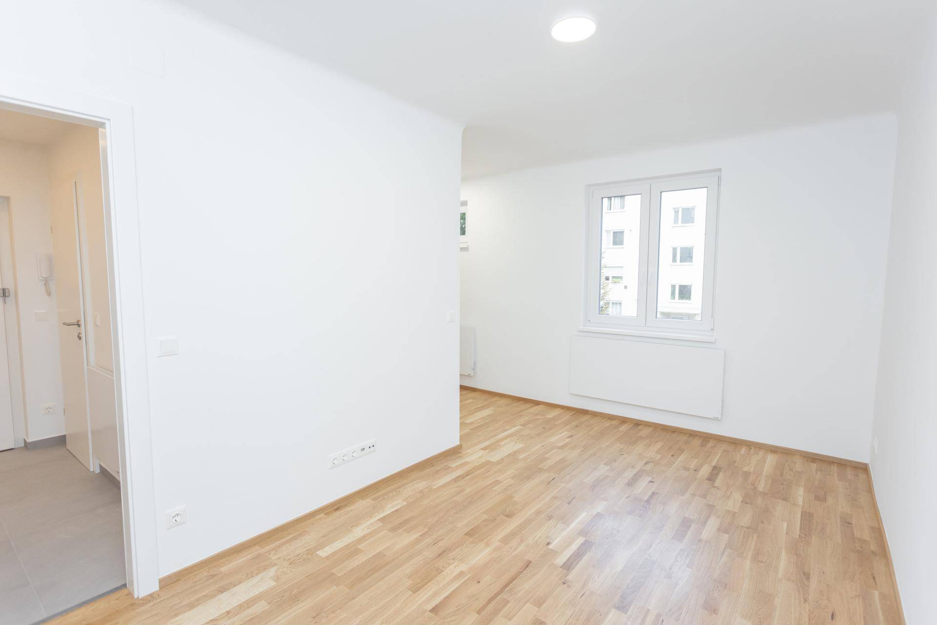 Heller, leerer Raum mit Holzfußboden und Fenster, angrenzend an einen gefliesten Flur.