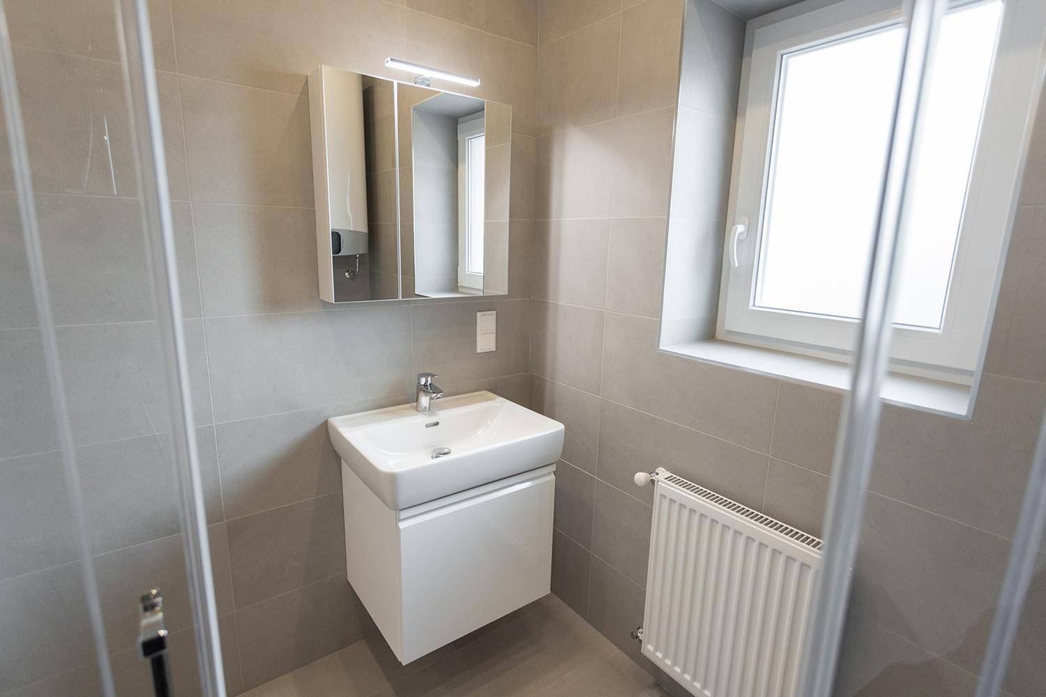 Helles Badezimmer mit grauen Fliesen, Waschbecken, Spiegelschrank und Dusche.