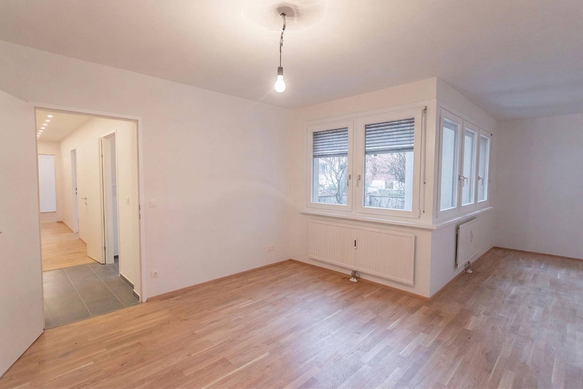 Heller, leerer Wohnraum mit Holzfußboden, weißen Wänden und einem Erkerfenster mit Jalousien.