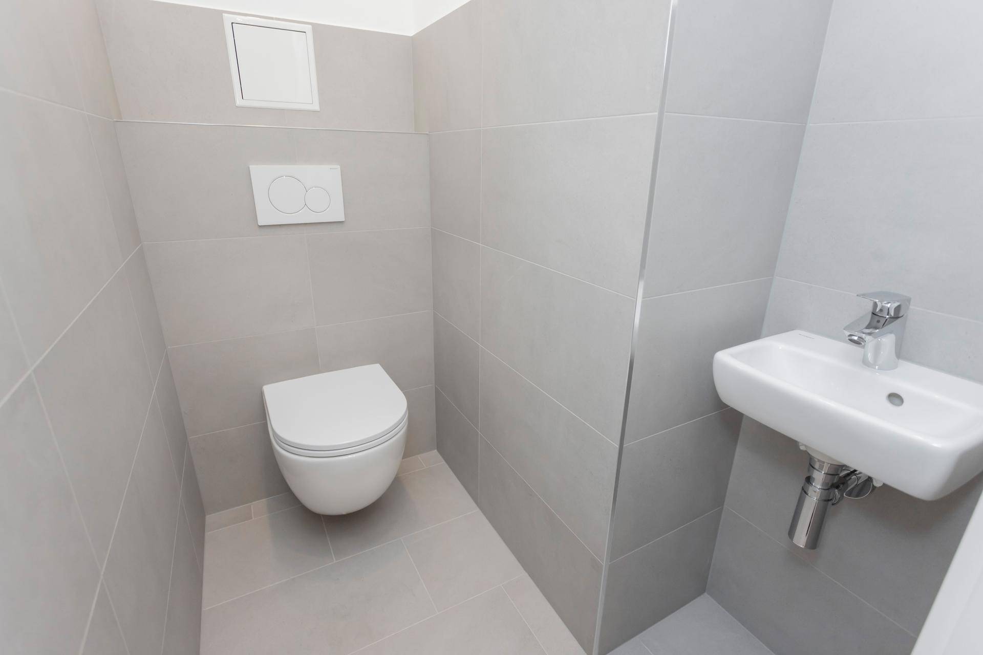 Modernes Gäste-WC mit wandhängender Toilette, kleinem Waschbecken und grauen Fliesen.