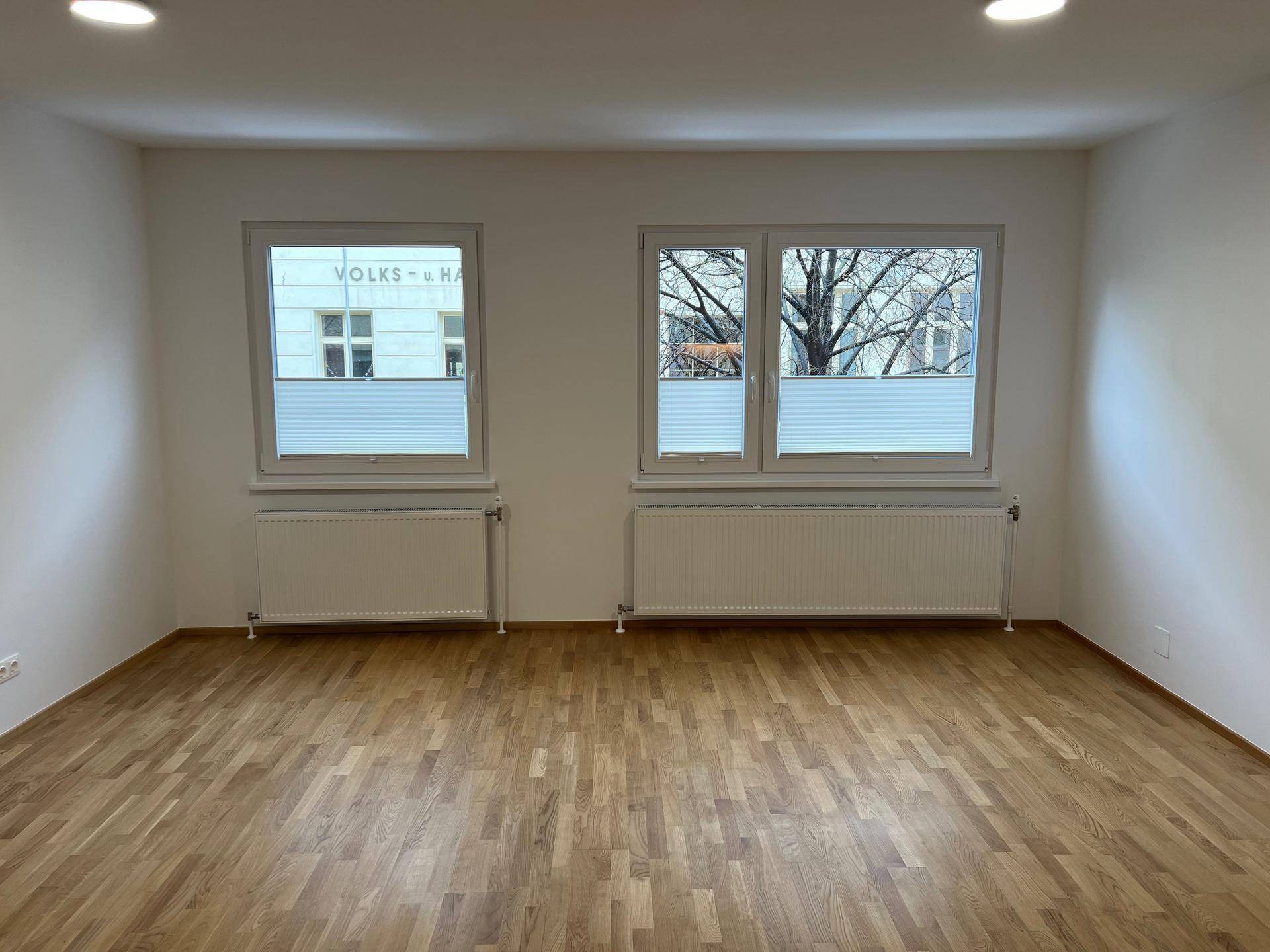 Helles Wohnzimmer mit Holzboden, zwei Fenstern und Deckenstrahlern, bereit zur Einrichtung.