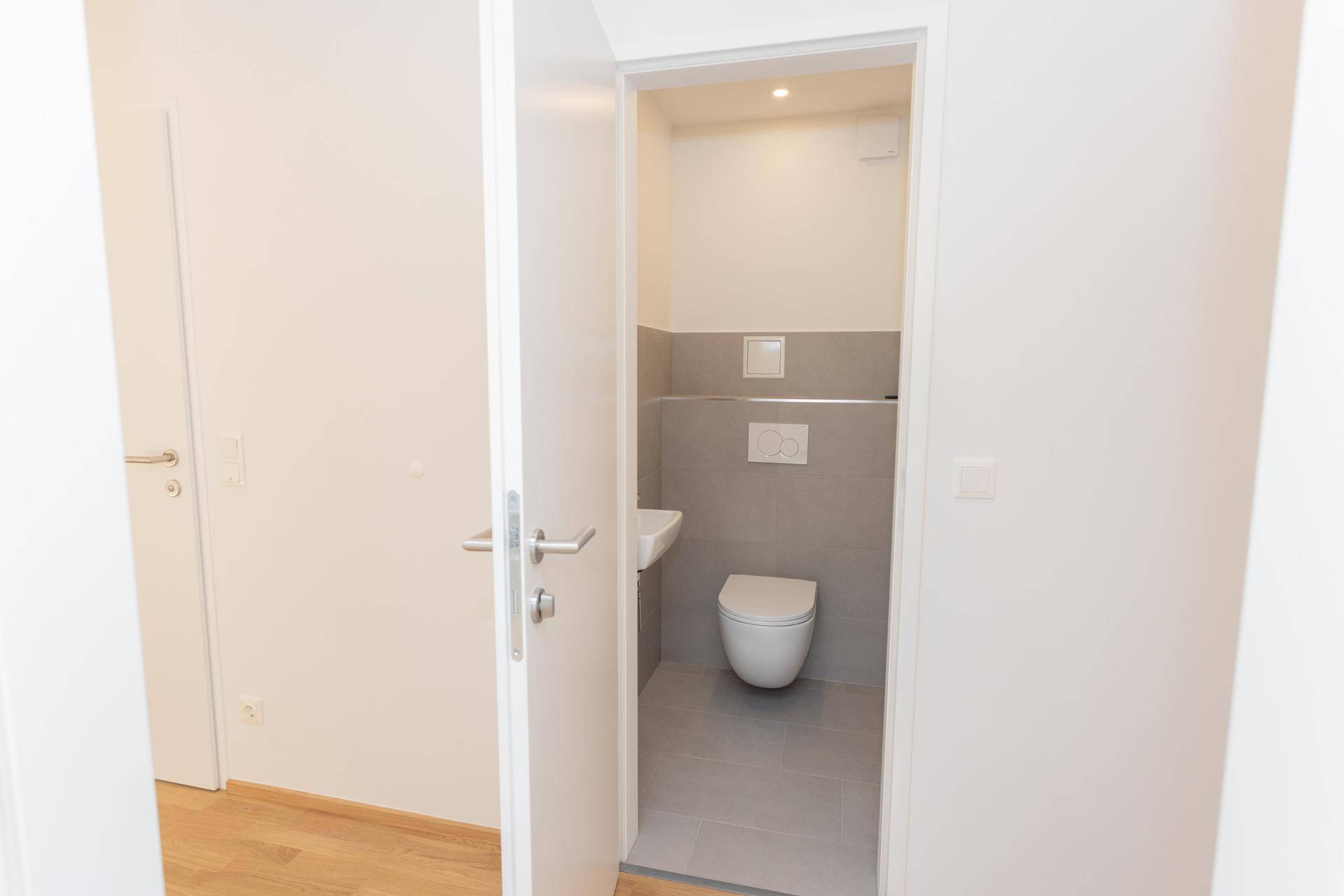 Modernes Gäste-WC mit Wand-WC, kleinem Waschbecken und grauen Fliesen, neu saniert.