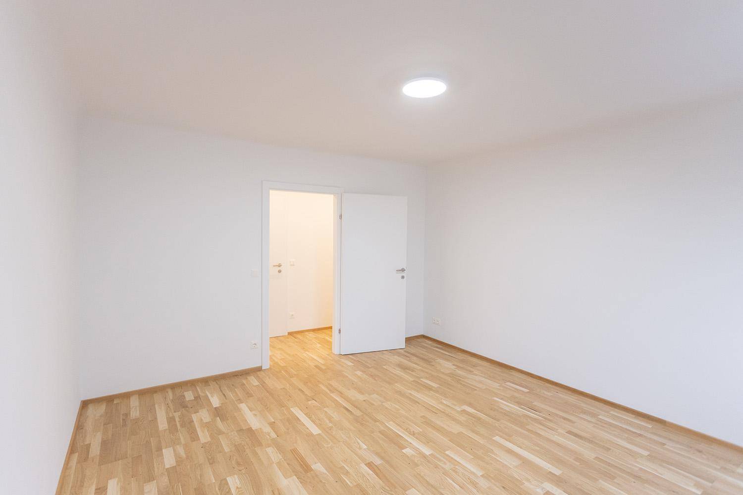 Helles Zimmer mit Holzboden und offener Tür, die den Zugang zu einem weiteren Raum ermöglicht.