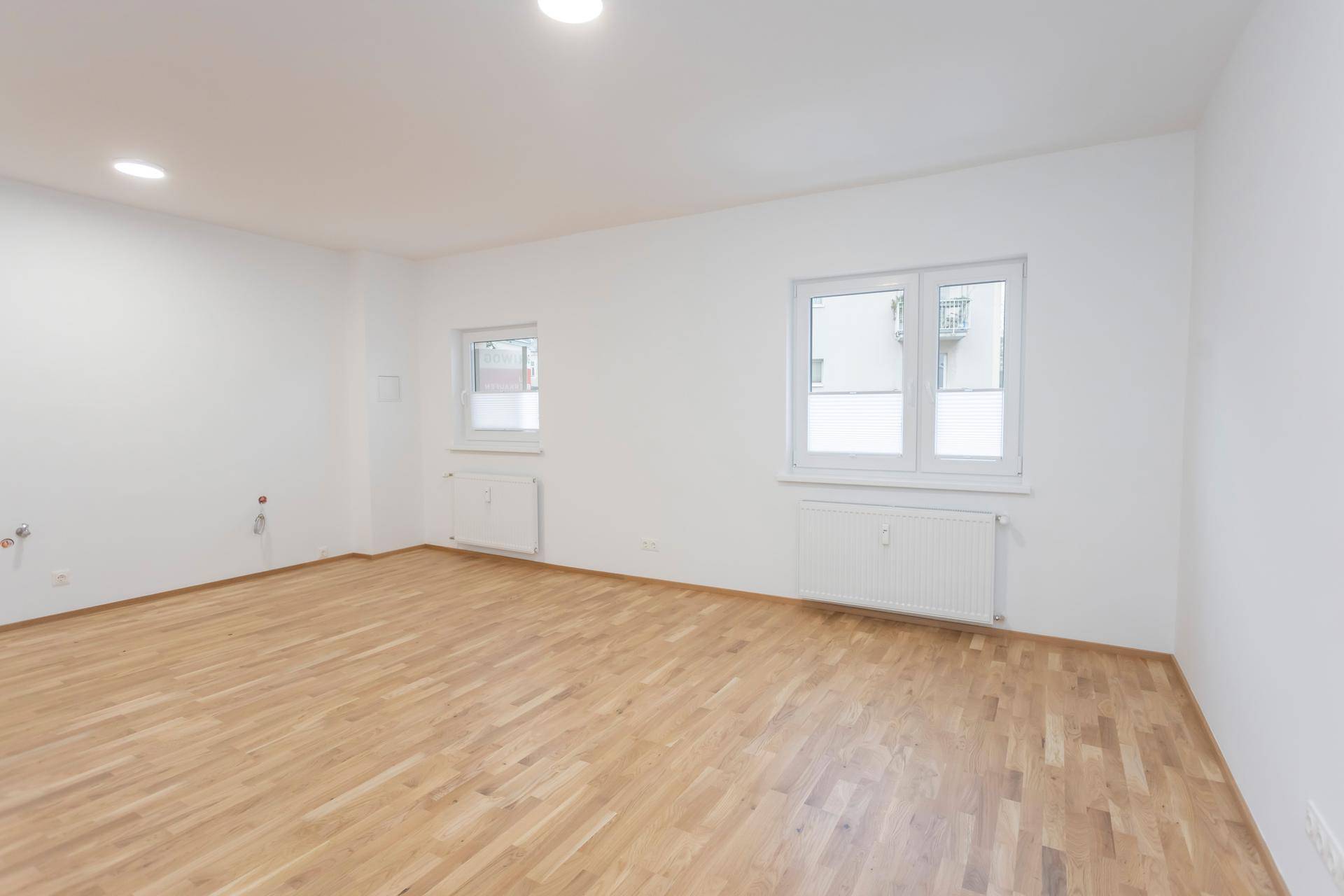 Helles, leeres Wohnzimmer mit Parkettboden und Blick nach draußen durch die Fenster.