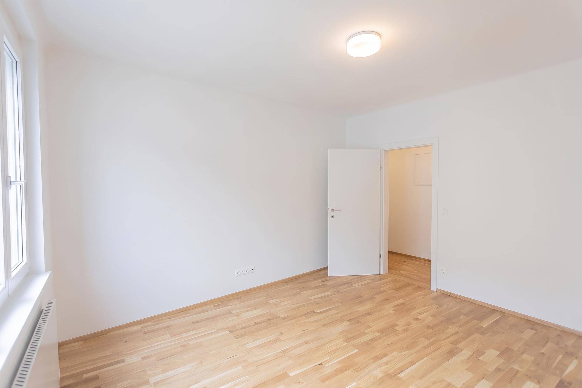Leerer Raum mit hellem Holzfußboden, weißen Wänden und Zugang zu einem angrenzenden Bereich.