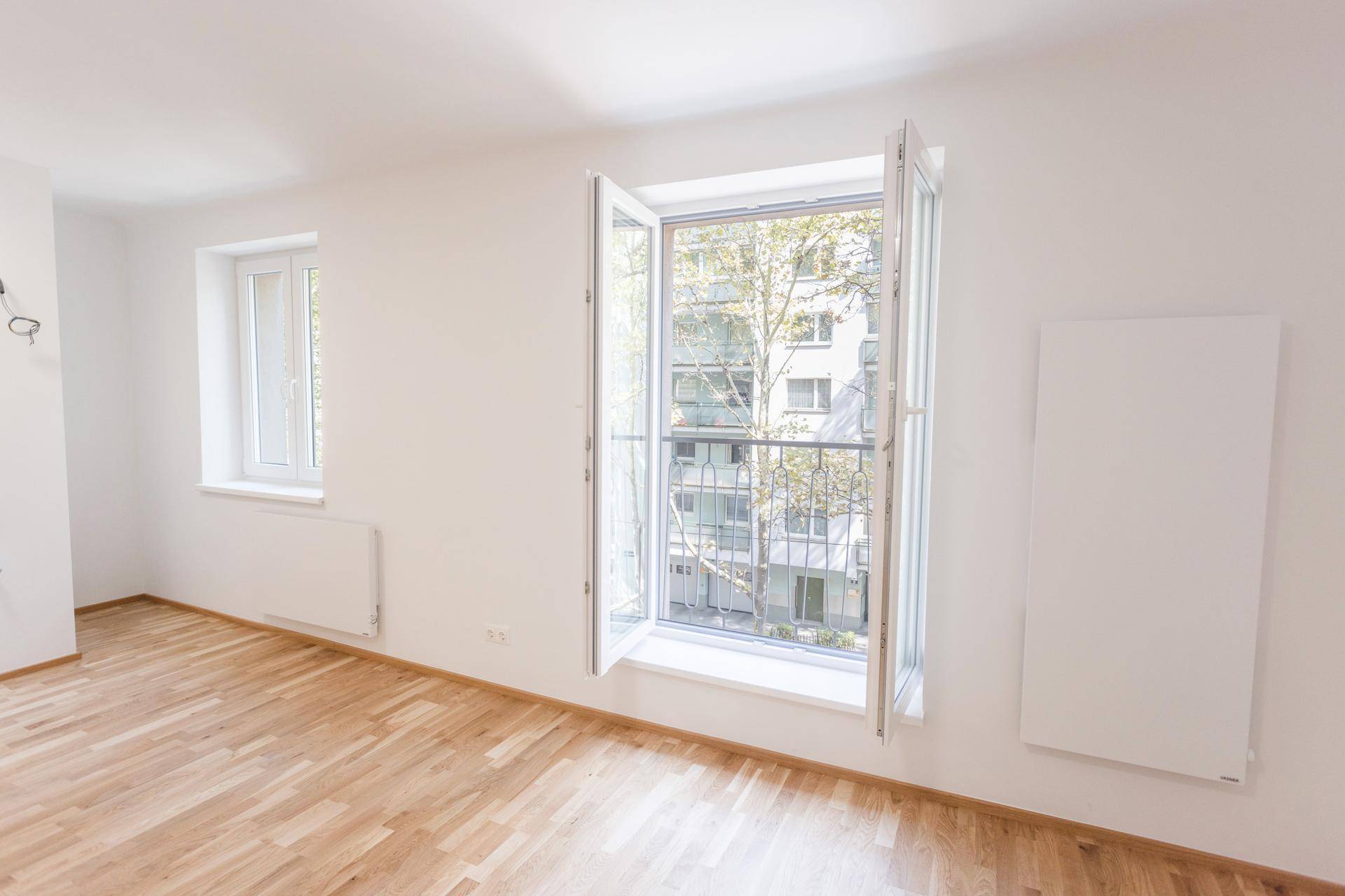 Wohnzimmer mit Parkettboden und geöffneten Fenstern, die einen grünen Ausblick bieten.