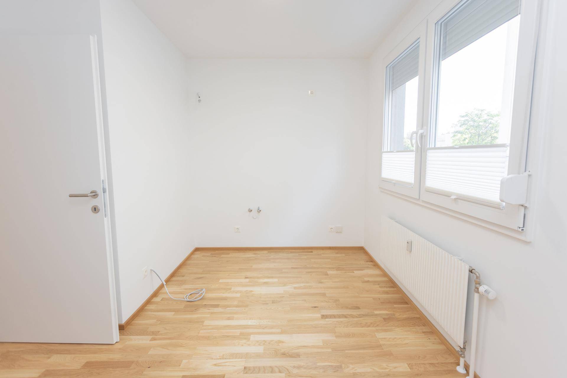 Heller, leerer Raum mit Parkettboden, großem Fenster mit Rollläden und Heizkörper.
