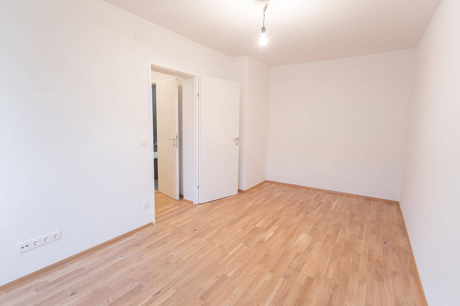 Unmöblierter Raum mit hellem Parkettboden und weiß gestrichenen Wänden, mit Zugang zu einem weiteren Zimmer.