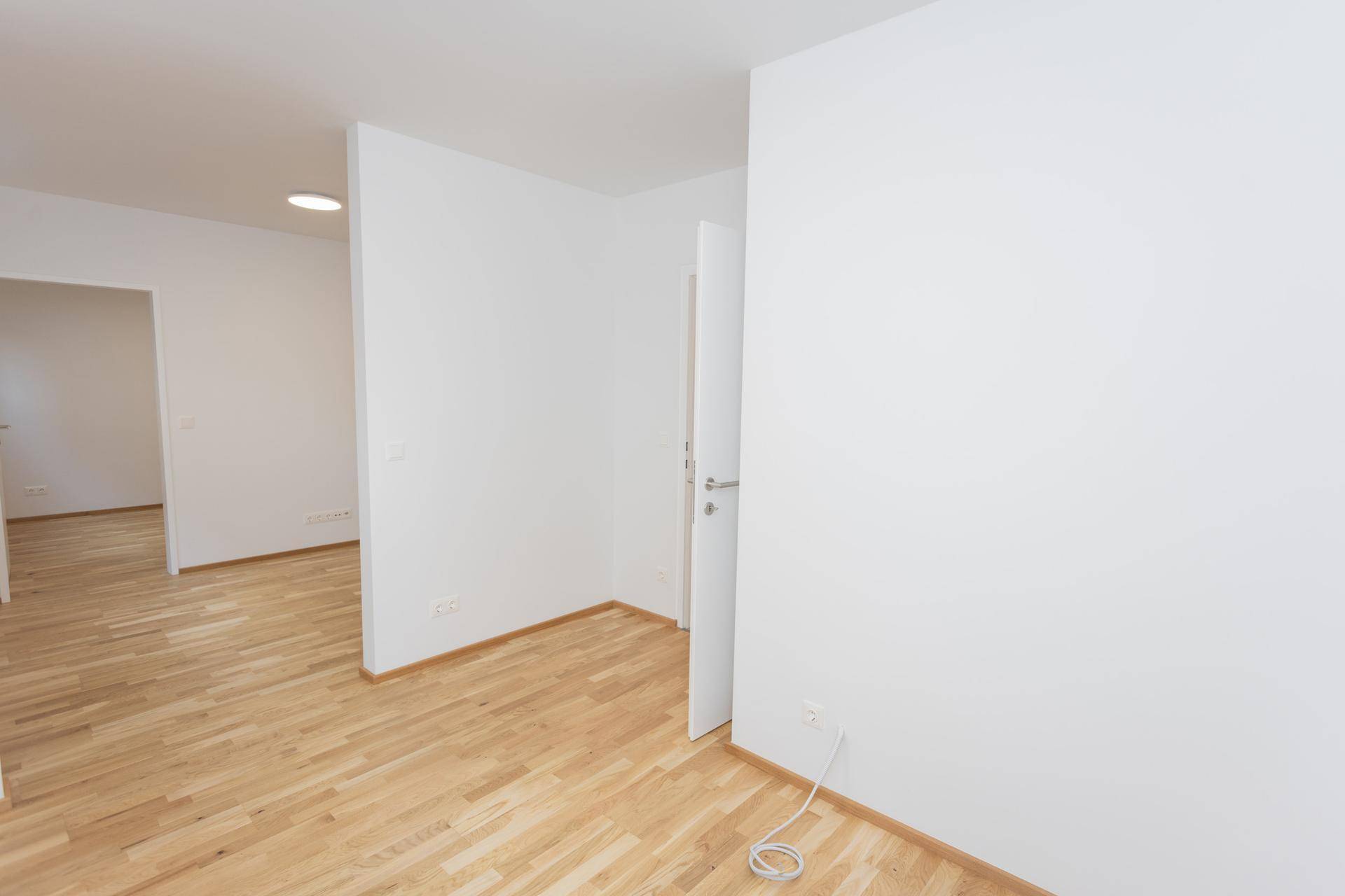Leerer Raum mit Parkettboden und weiß gestrichenen Wänden, mit Blick in den Flur.