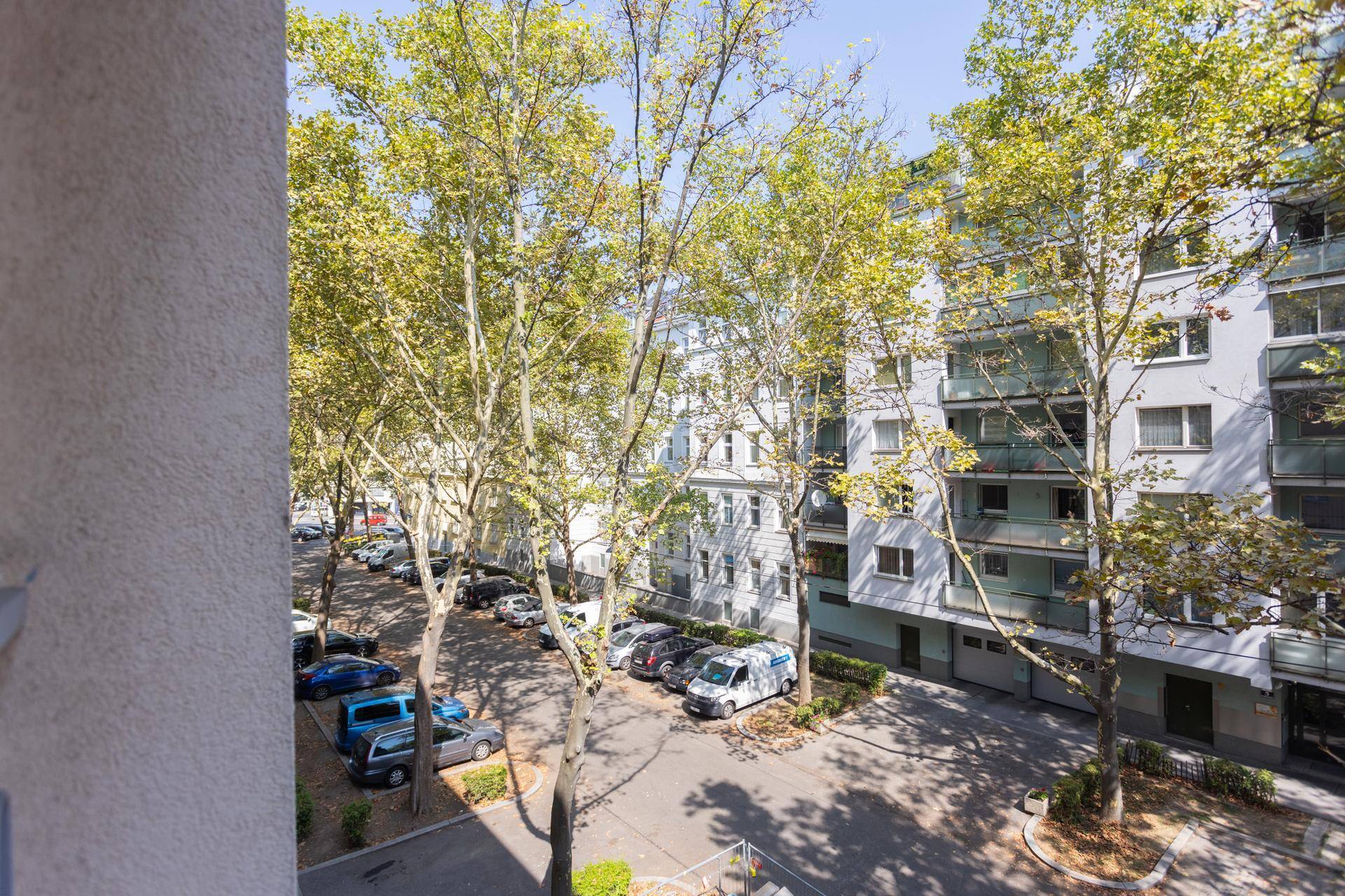 Blick vom Balkon auf die Straße mit parkenden Autos und Bäumen vor Wohngebäuden.