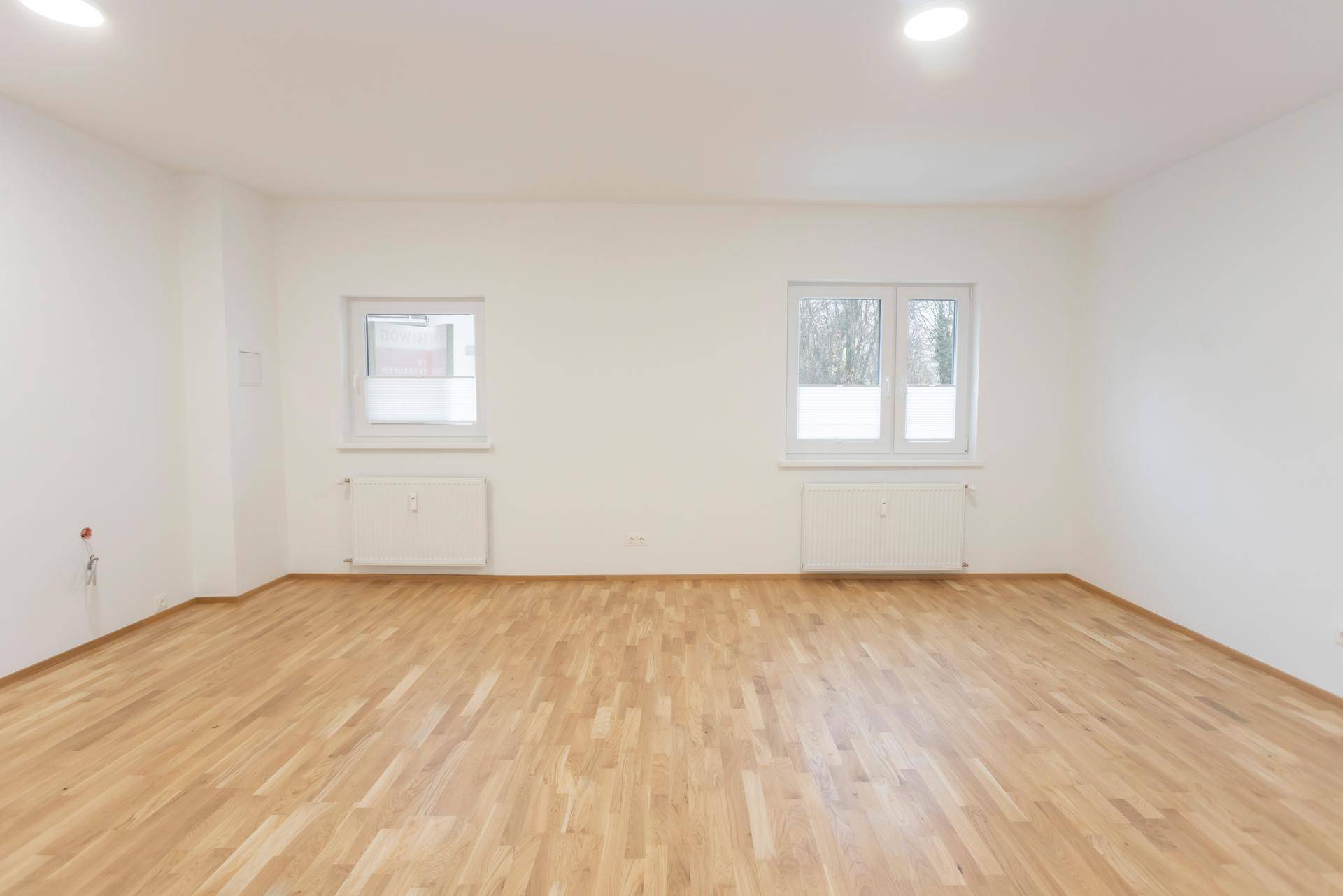 Geräumiges, leeres Wohnzimmer mit hellem Parkettboden und zwei Fenstern.