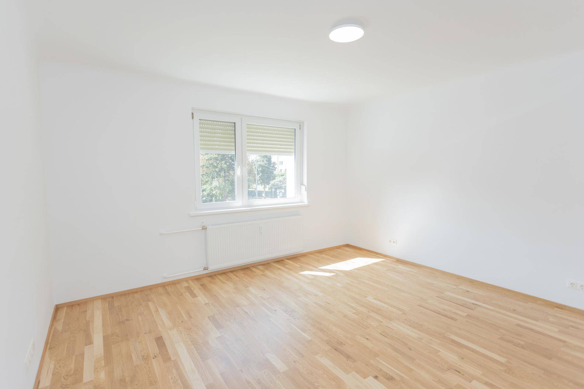 Leeres, helles Schlafzimmer mit Holzboden und großem Fenster, das viel Tageslicht hereinlässt.