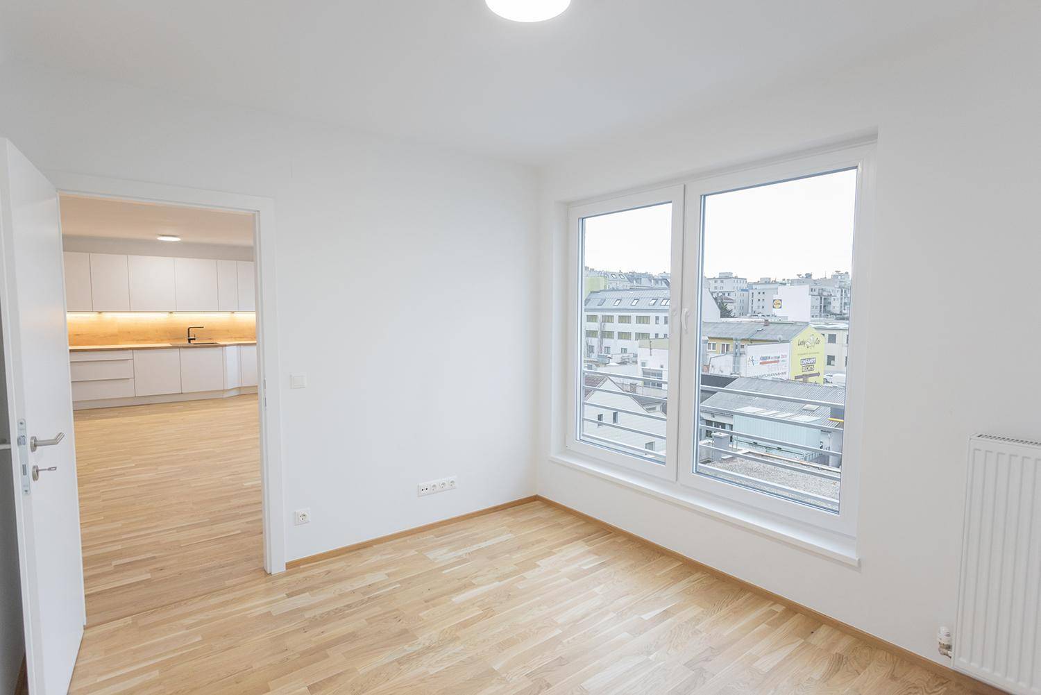 Heller Raum mit Holzboden und Blick auf die umliegenden Gebäude, mit Zugang zur Küche.