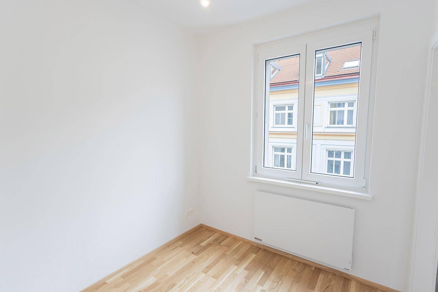 Leeres Zimmer mit hellem Parkettboden und einem Fenster, das auf ein Wohngebäude blickt.