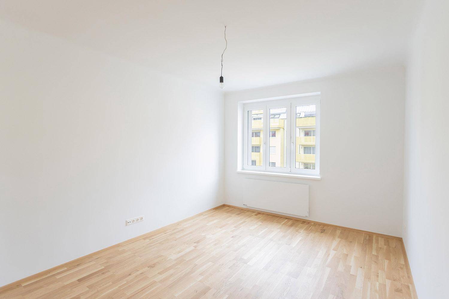 Helles, leeres Zimmer mit Parkettboden und einem Fenster, das auf ein Wohngebäude blickt.