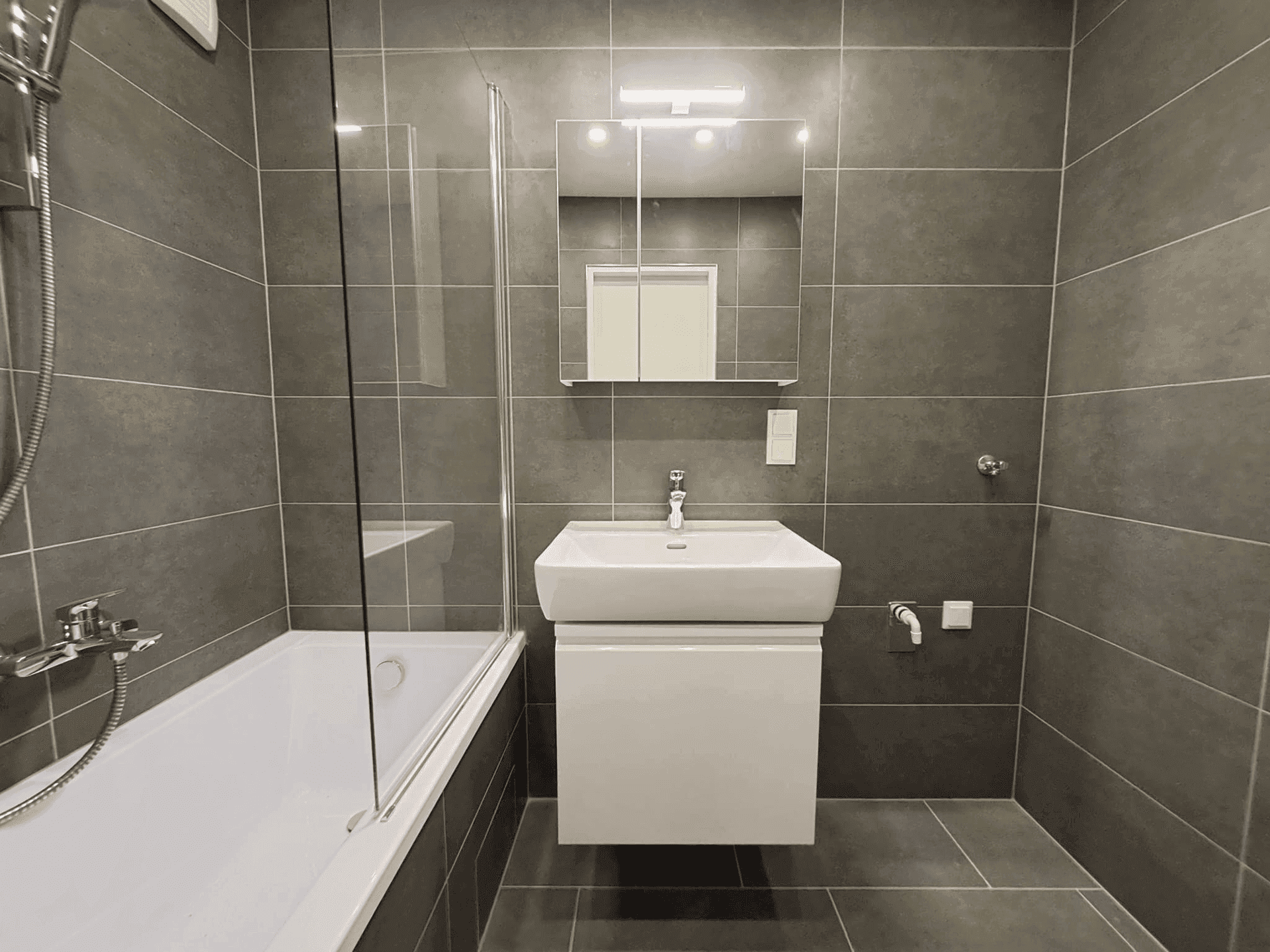 Funktionales Badezimmer mit grauen Fliesen, Badewanne, Waschtischunterschrank und Spiegelschrank.