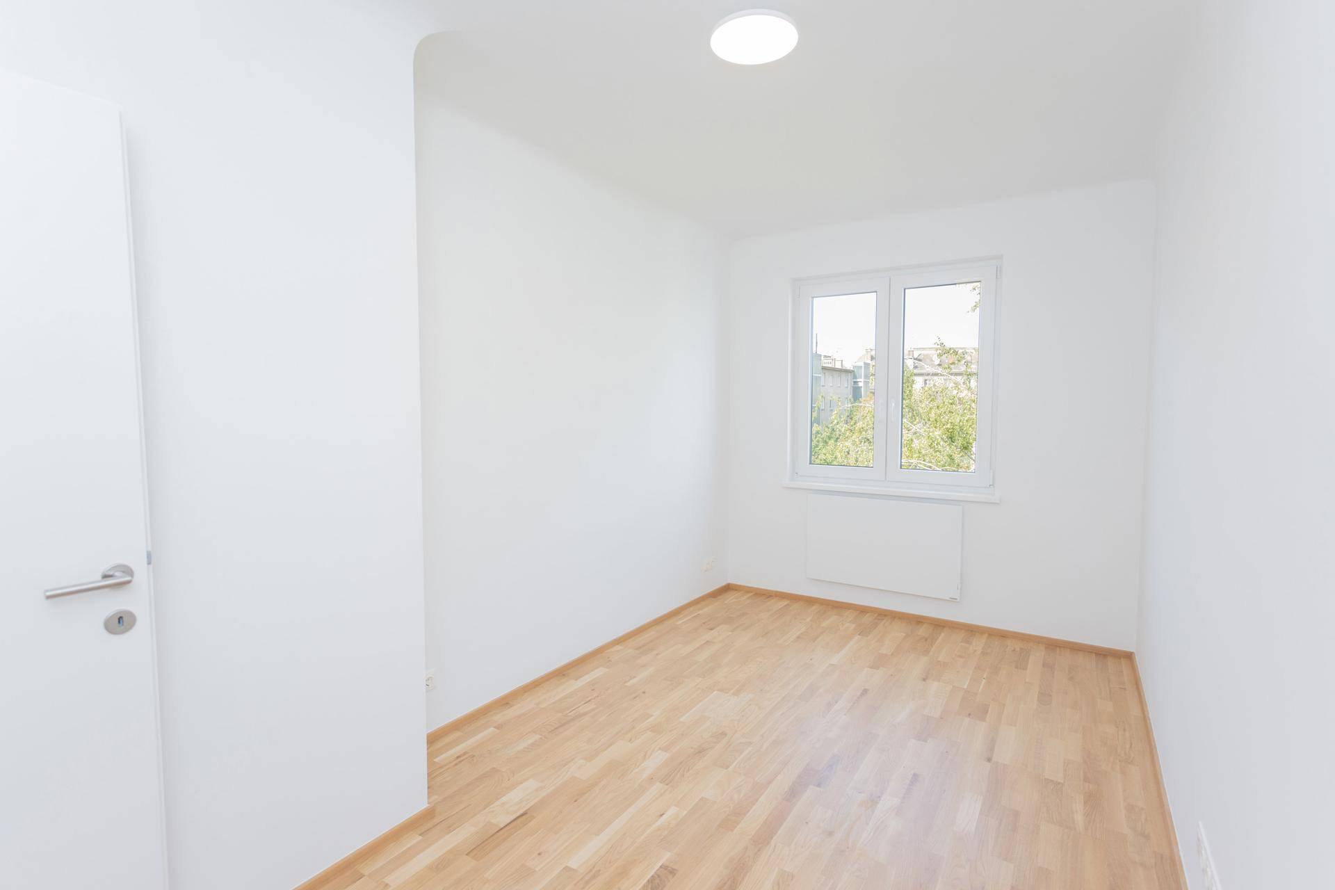 Leeres Schlafzimmer mit hellem Parkettboden und einem Fenster, das den Raum erhellt.