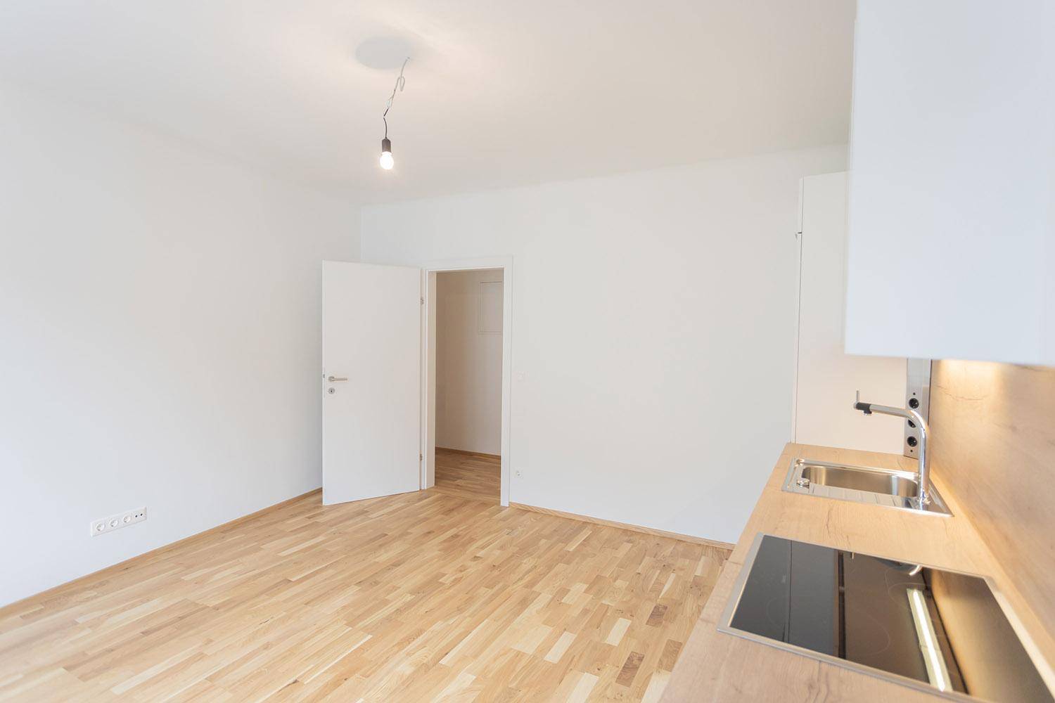 Heller, leerer Wohnbereich mit Holzboden und Blick auf die moderne Küchenzeile.