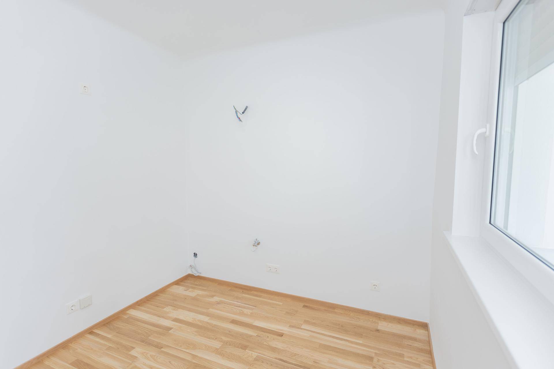 Helles, leeres Zimmer mit Parkettboden und einem Fenster, das viel Tageslicht hereinlässt.