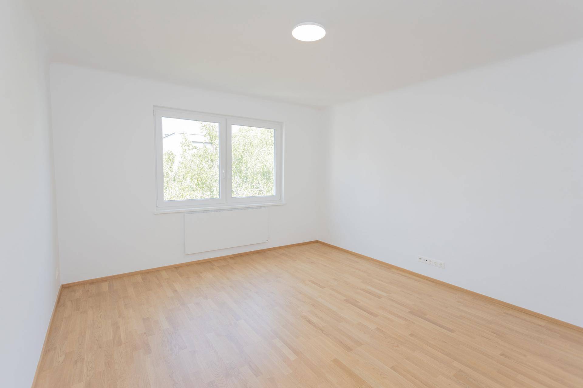 Helles, leeres Schlafzimmer mit Parkettboden und Fenster, das viel Tageslicht hereinlässt.