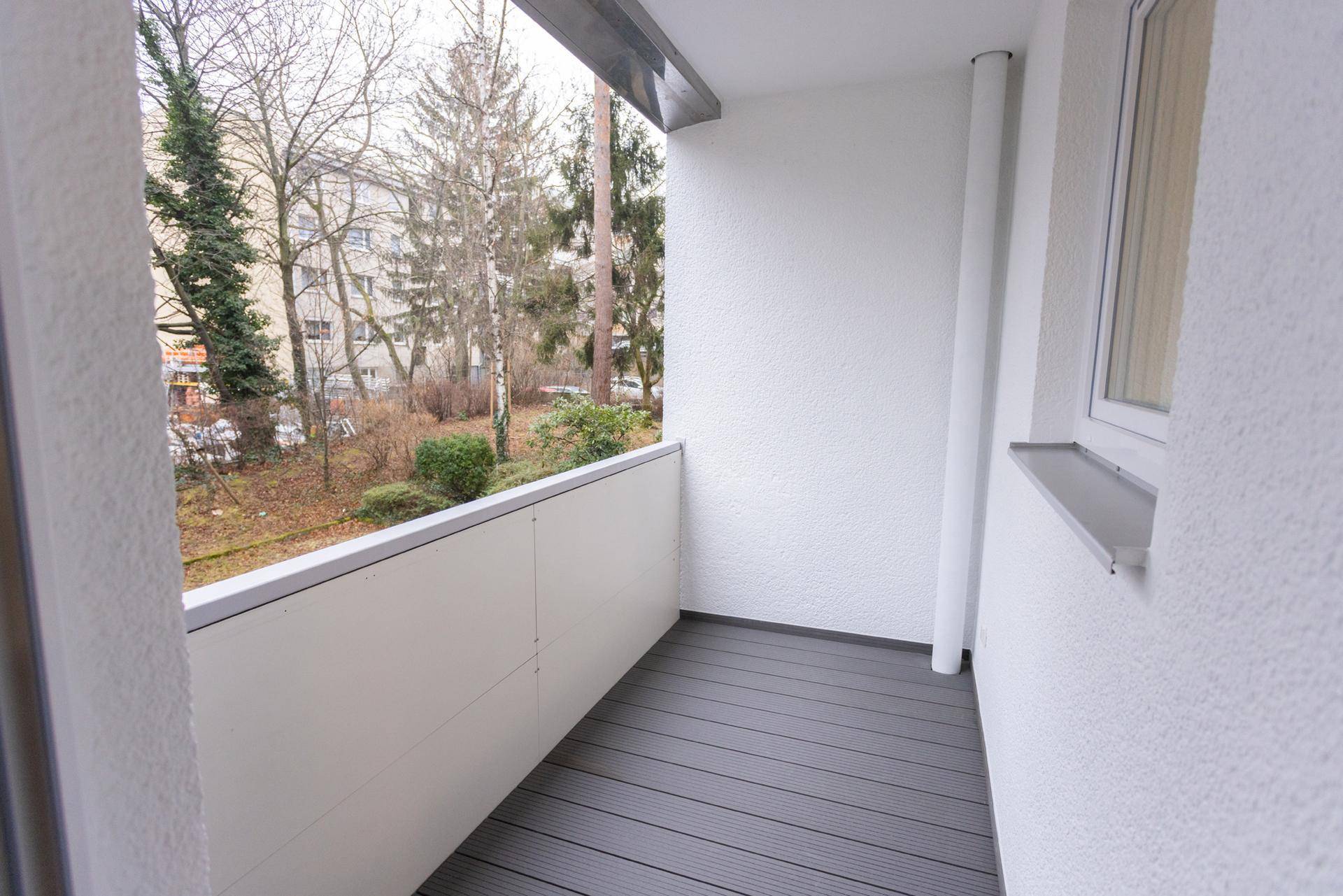 Leerer Balkon mit grauem Bodenbelag und Blick auf Bäume und umliegende Gebäude.