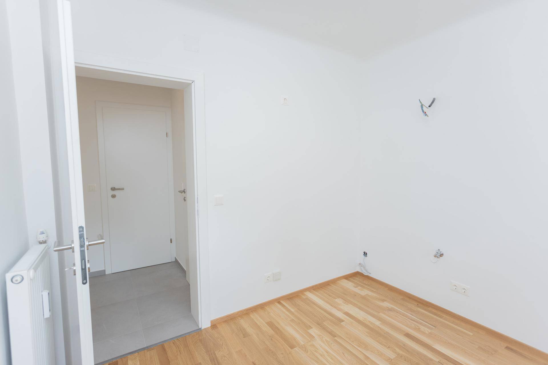 Leeres Zimmer mit hellem Parkettboden, weiß gestrichenen Wänden und Zugang zu einem Flur.