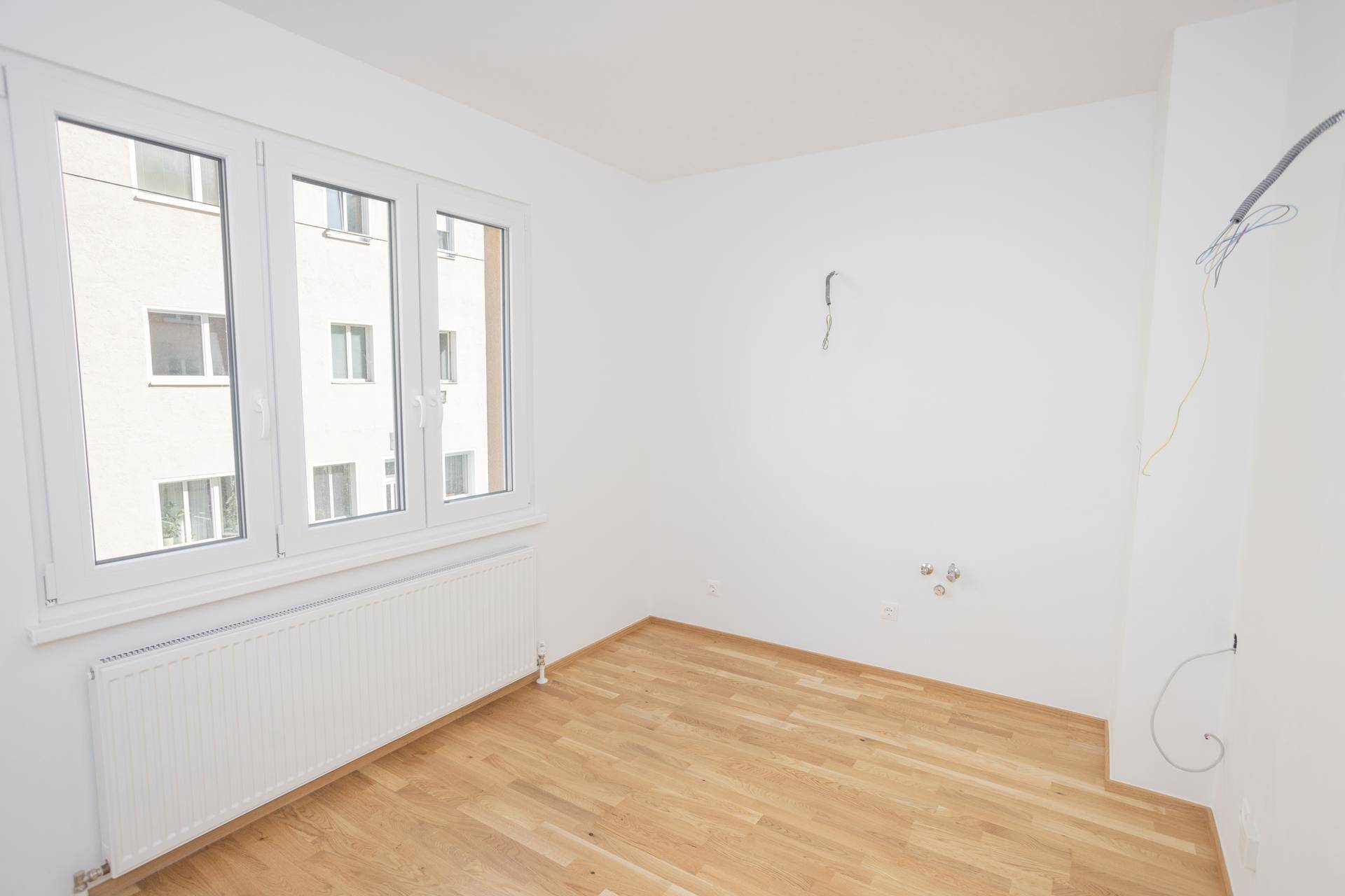 Leeres Zimmer mit hellem Parkettboden, weißen Wänden und einem großen Fenster mit Heizkörper.