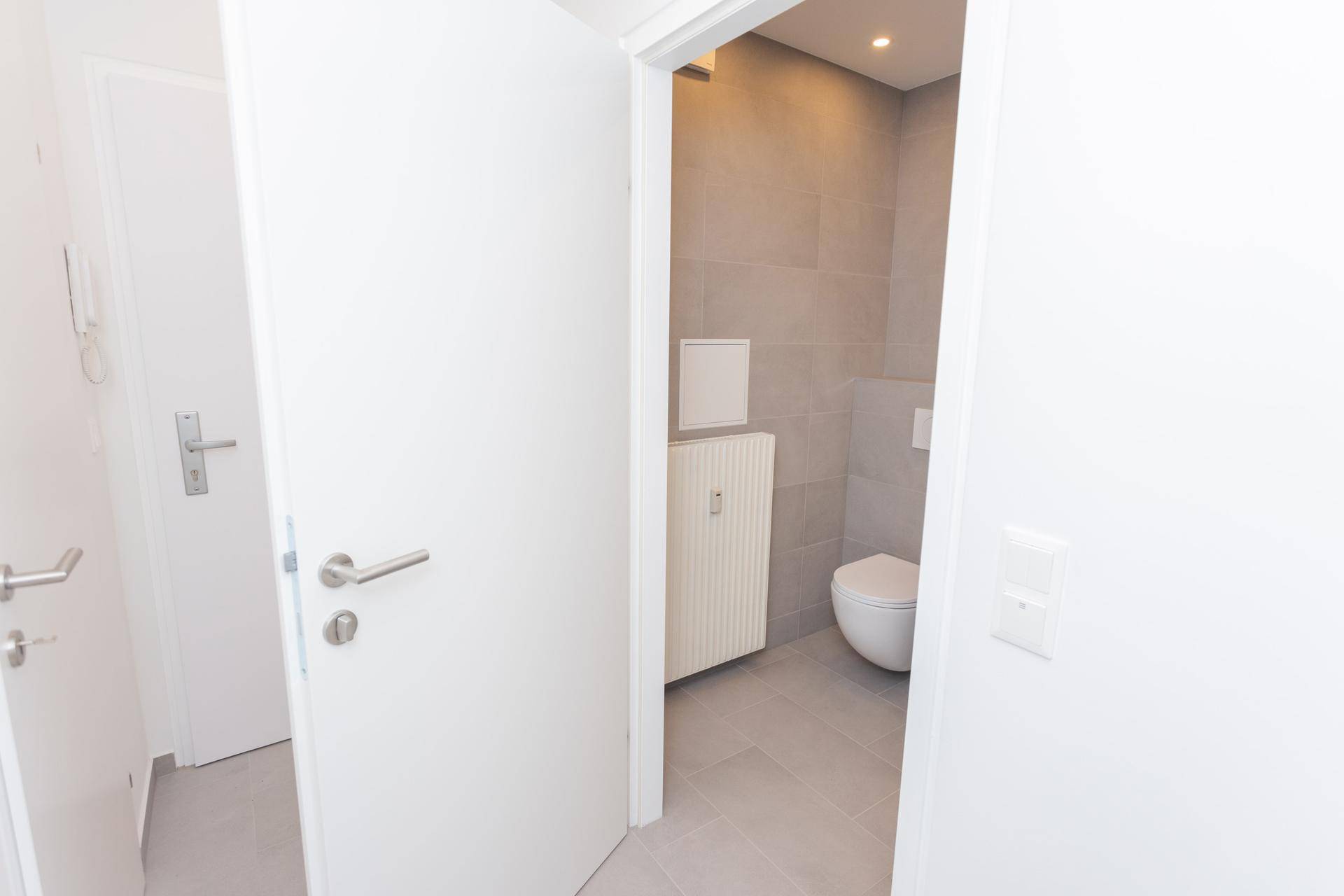 Blick vom Flur in ein modernes WC mit grauen Fliesen und wandhängender Toilette.