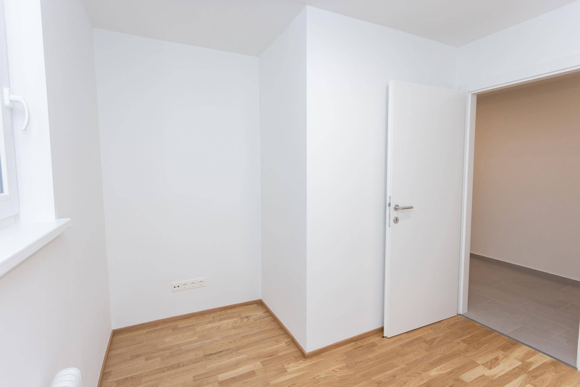 Heller, leerer Raum mit Parkettboden und offener Tür zu einem gefliesten Bereich.