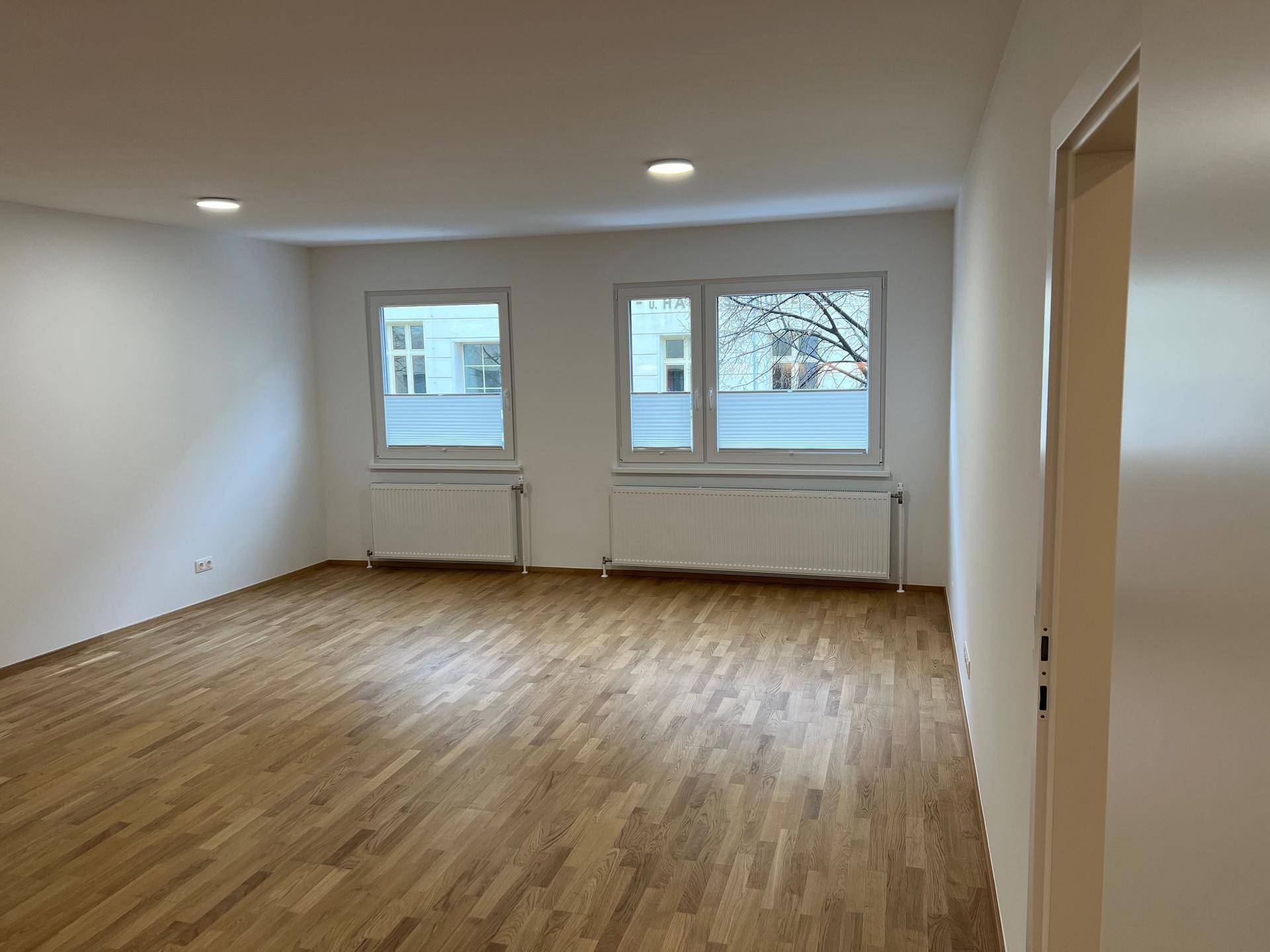 Helles Wohnzimmer mit Holzboden, zwei Fenstern und Deckenstrahlern, bereit zur Einrichtung.
