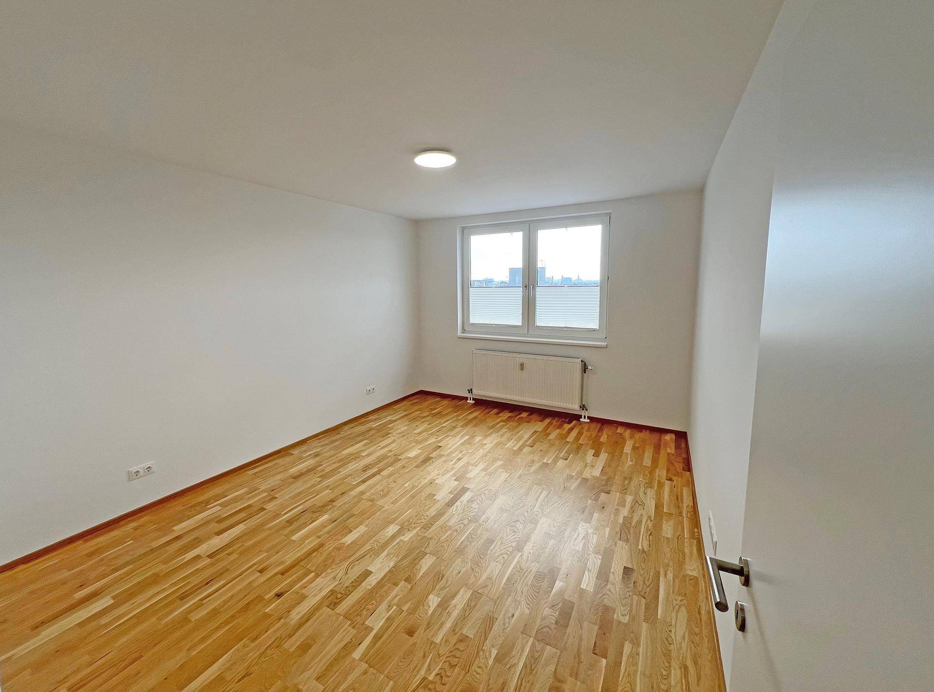 Helles, leeres Zimmer mit Parkettboden und Fenster mit Blick auf die Stadt.