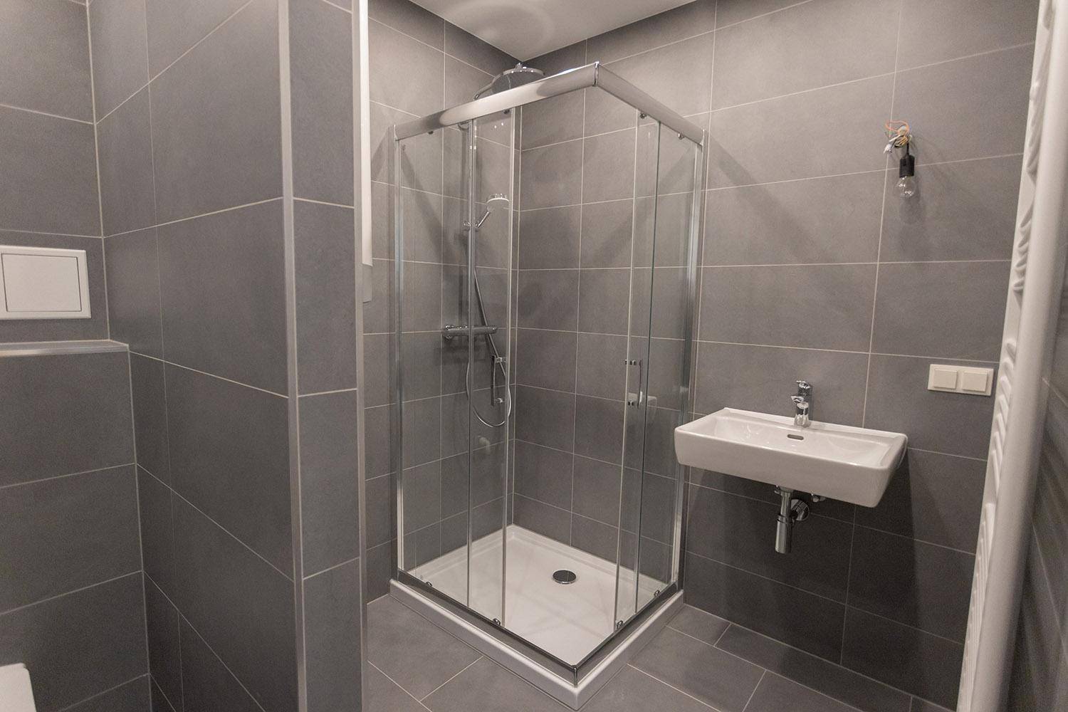 Duschkabine und kleines Waschbecken in einem modern gefliesten Badezimmer mit grauen Wand- und Bodenfliesen.