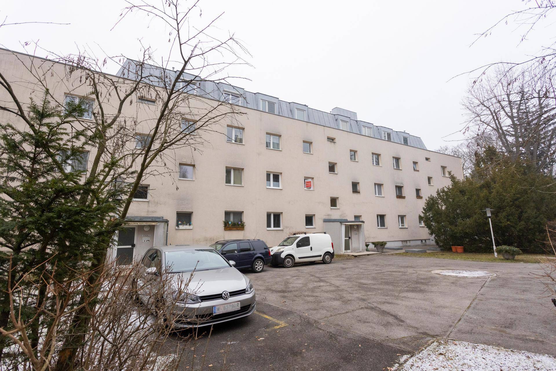 Mehrfamilienhaus mit Parkplätzen im Freien und Bäumen im Vordergrund.