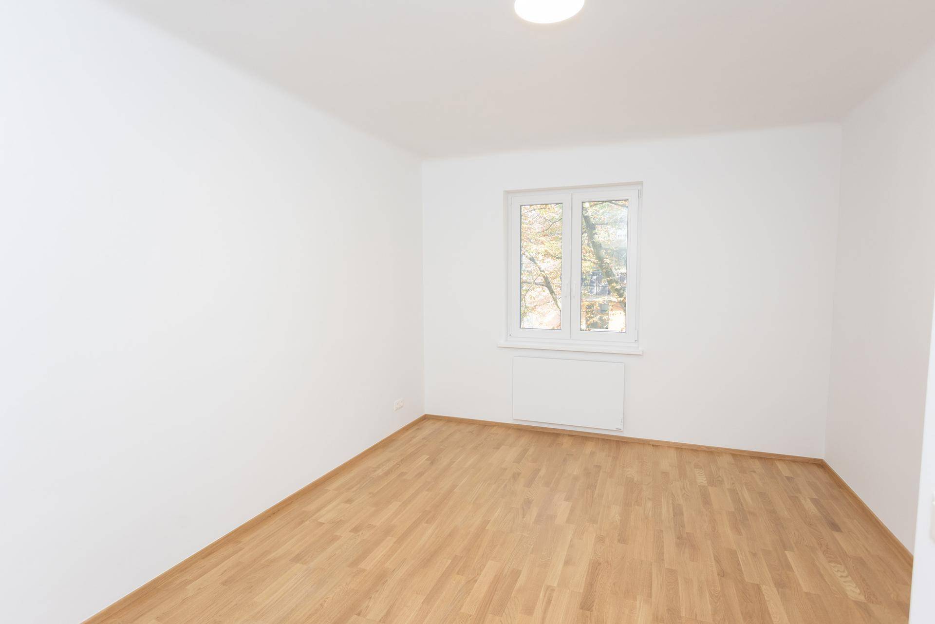 Leeres, helles Zimmer mit Holzparkettboden und einem großen Fenster mit Blick ins Grüne.