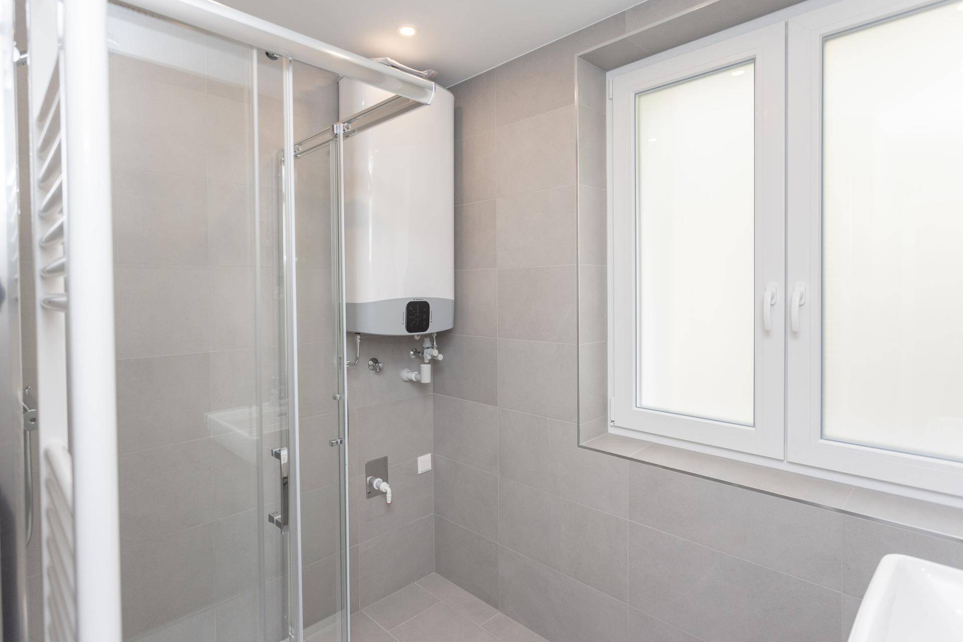 Modernes Badezimmer mit Duschkabine, Boiler und Fenster, komplett gefliest in Grau.