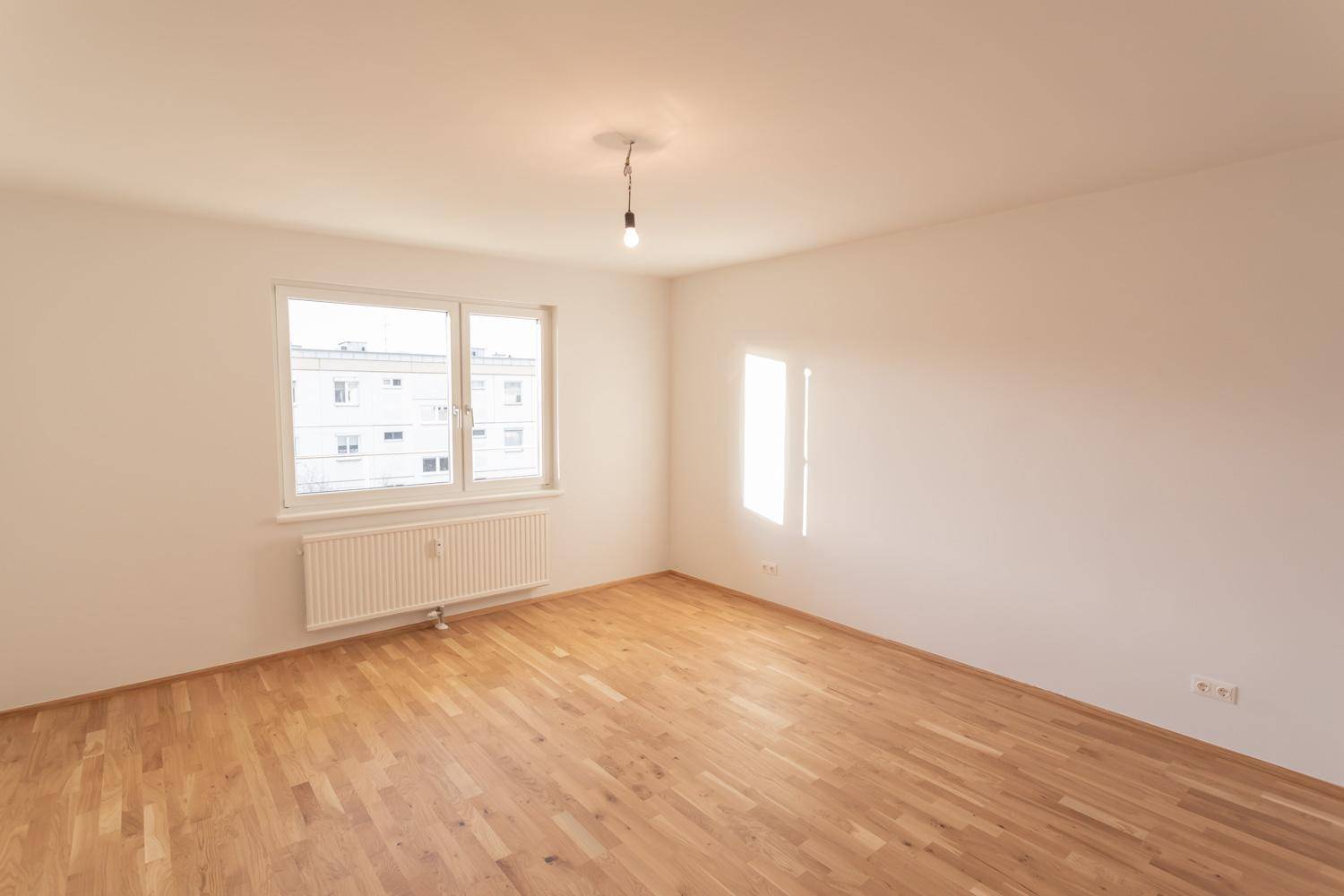 Helles, leeres Zimmer mit Parkettboden und großem Fenster für viel Tageslicht.