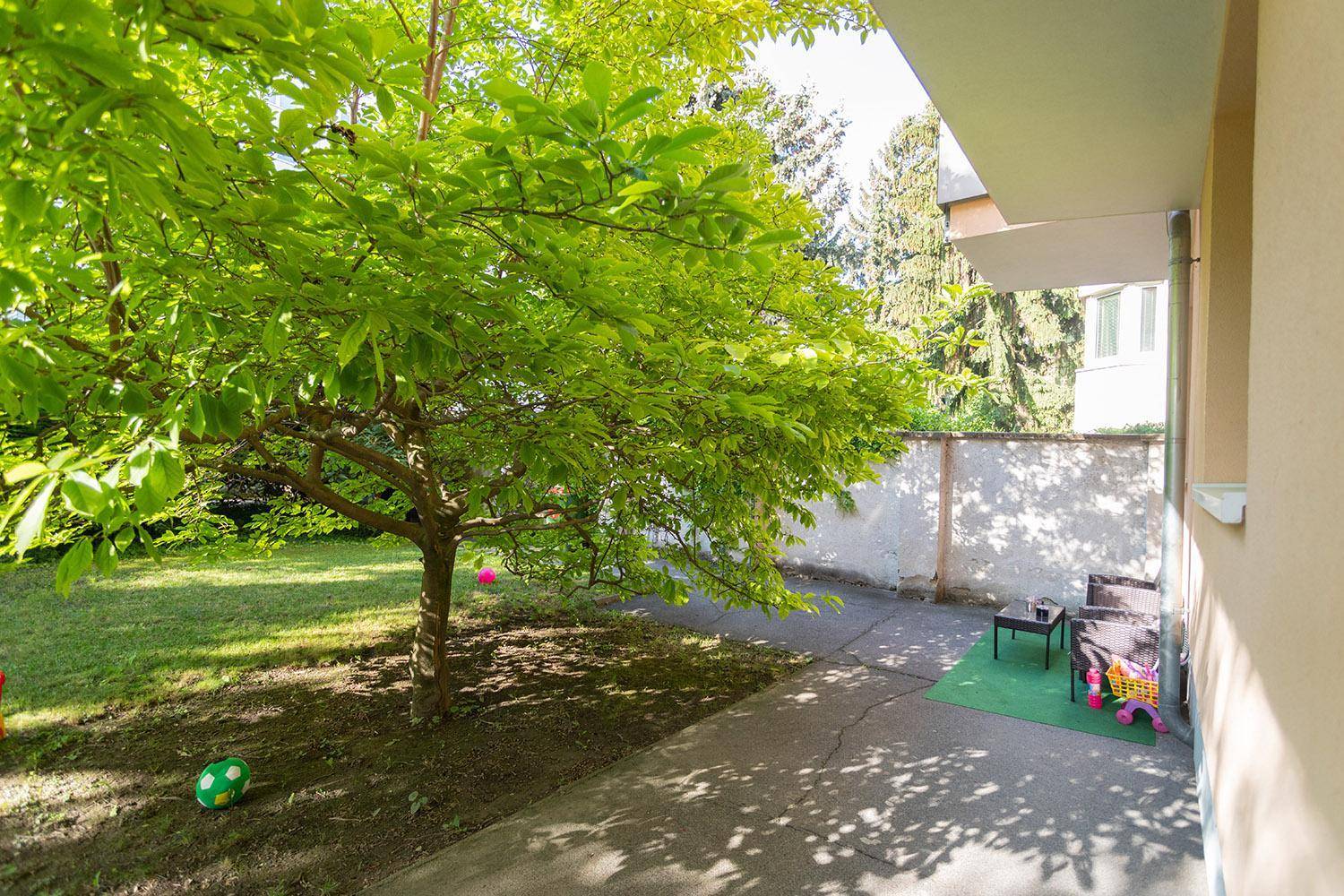Grüner Gartenbereich mit Baum und kleiner gepflasterter Fläche für Sitzgelegenheiten.