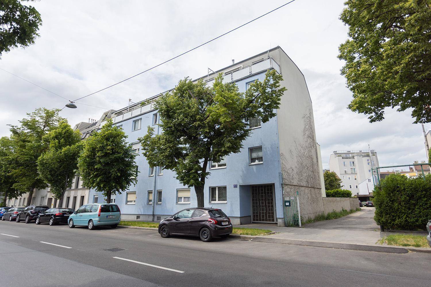 Mehrfamilienhaus mit blauer Fassade, Bäumen und parkenden Autos an der Straße.