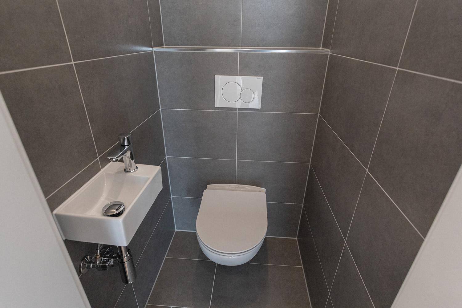 Modernes Gäste-WC mit grauen Fliesen, wandhängendem WC und kleinem Waschbecken.
