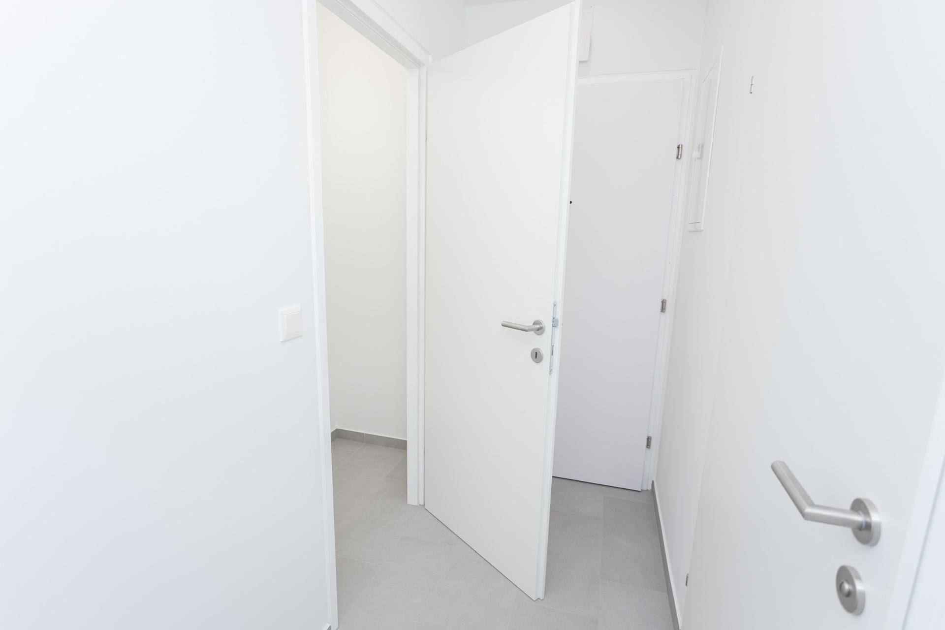 Weißer Flur mit offener Tür, die den Blick in einen weiteren Raum mit Fliesenboden freigibt.