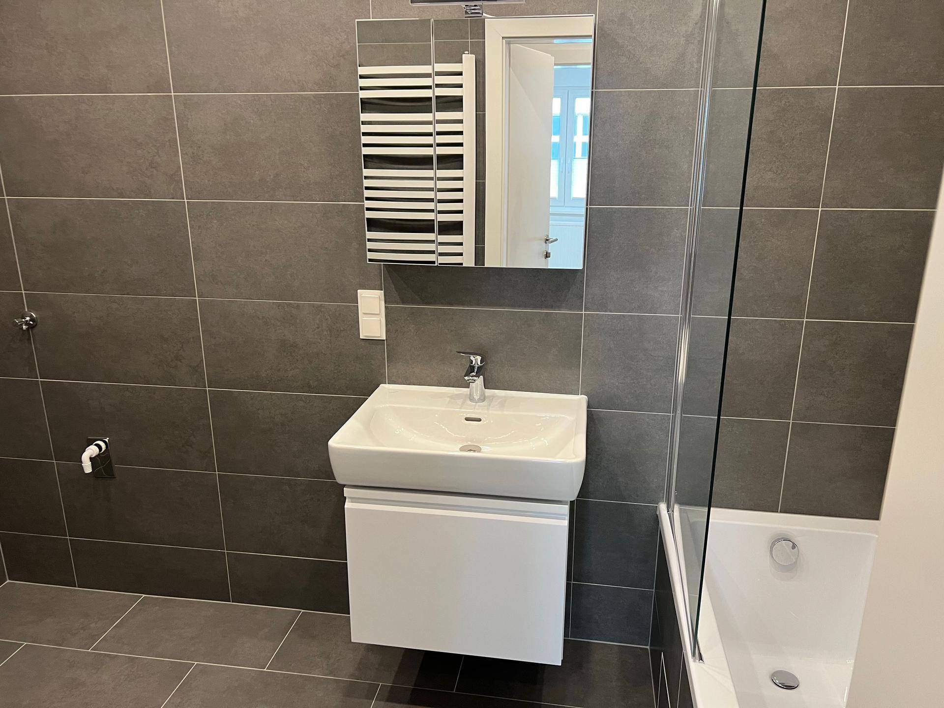 Modernes Badezimmer mit grauen Fliesen, wandhängendem Waschtisch, Spiegelschrank und Handtuchheizkörper.