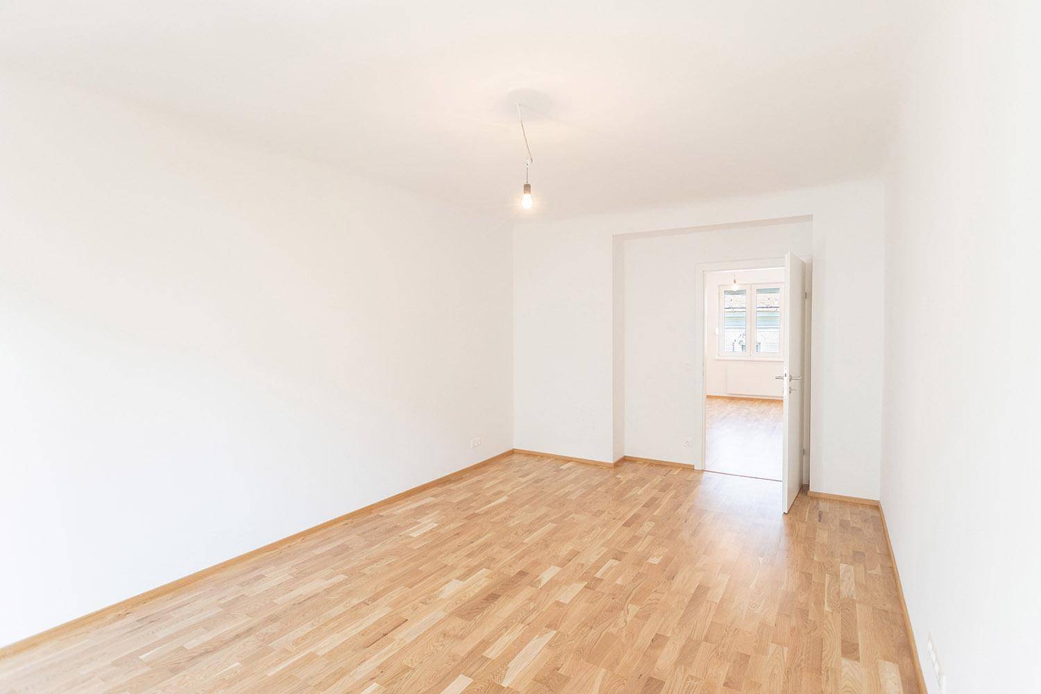 Heller, leerer Raum mit Parkettboden und offener Tür zu einem weiteren Zimmer.