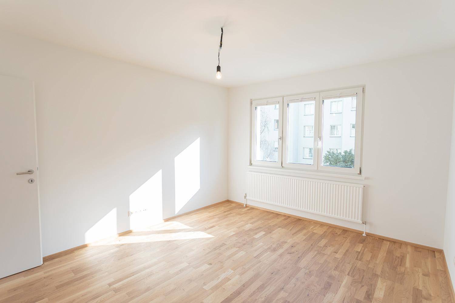 Leerer Raum mit hellem Parkettboden und Fenster, das viel Tageslicht hereinlässt.
