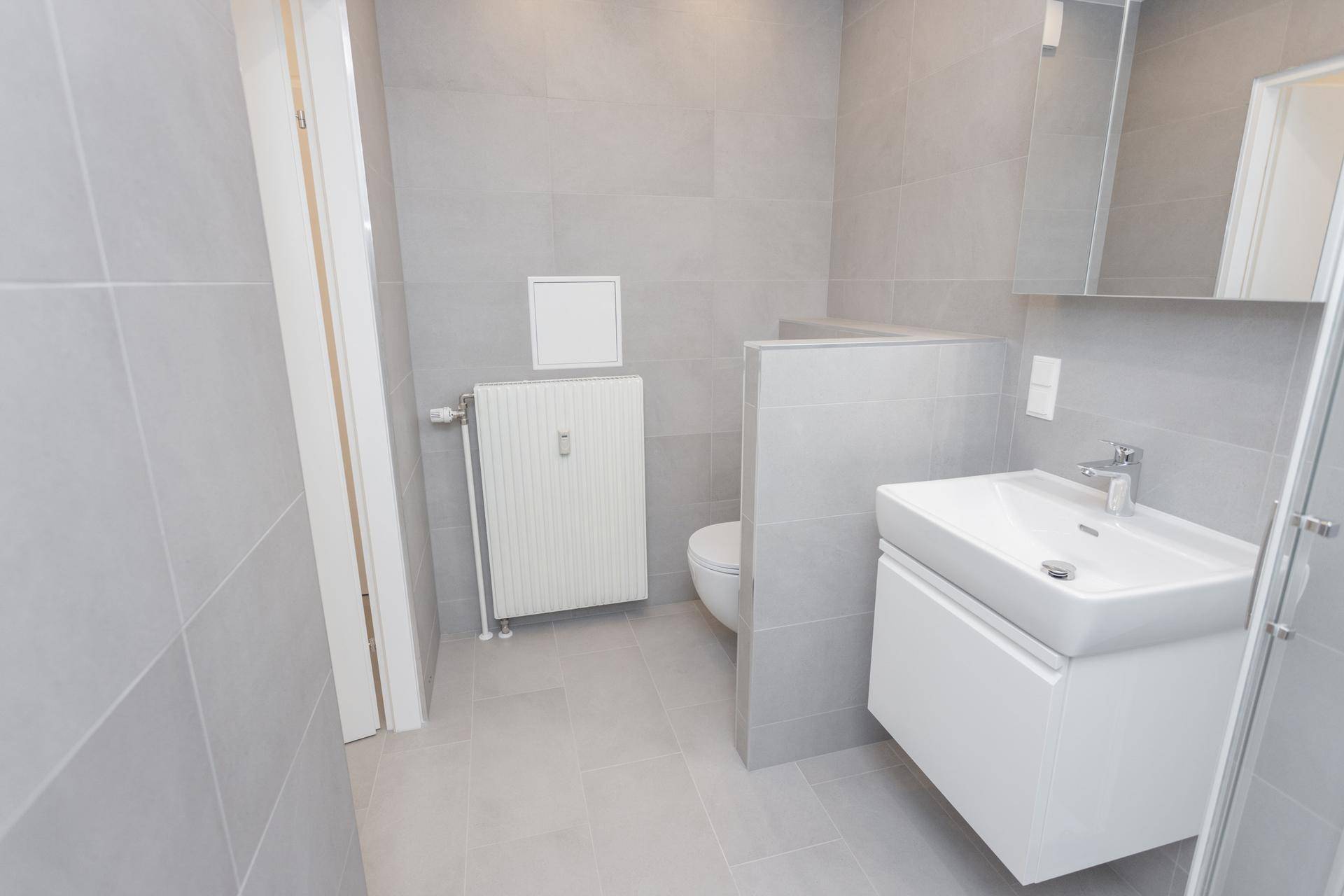 Modernes Badezimmer mit grauen Fliesen, Heizkörper, wandhängender Toilette und Waschbecken.
