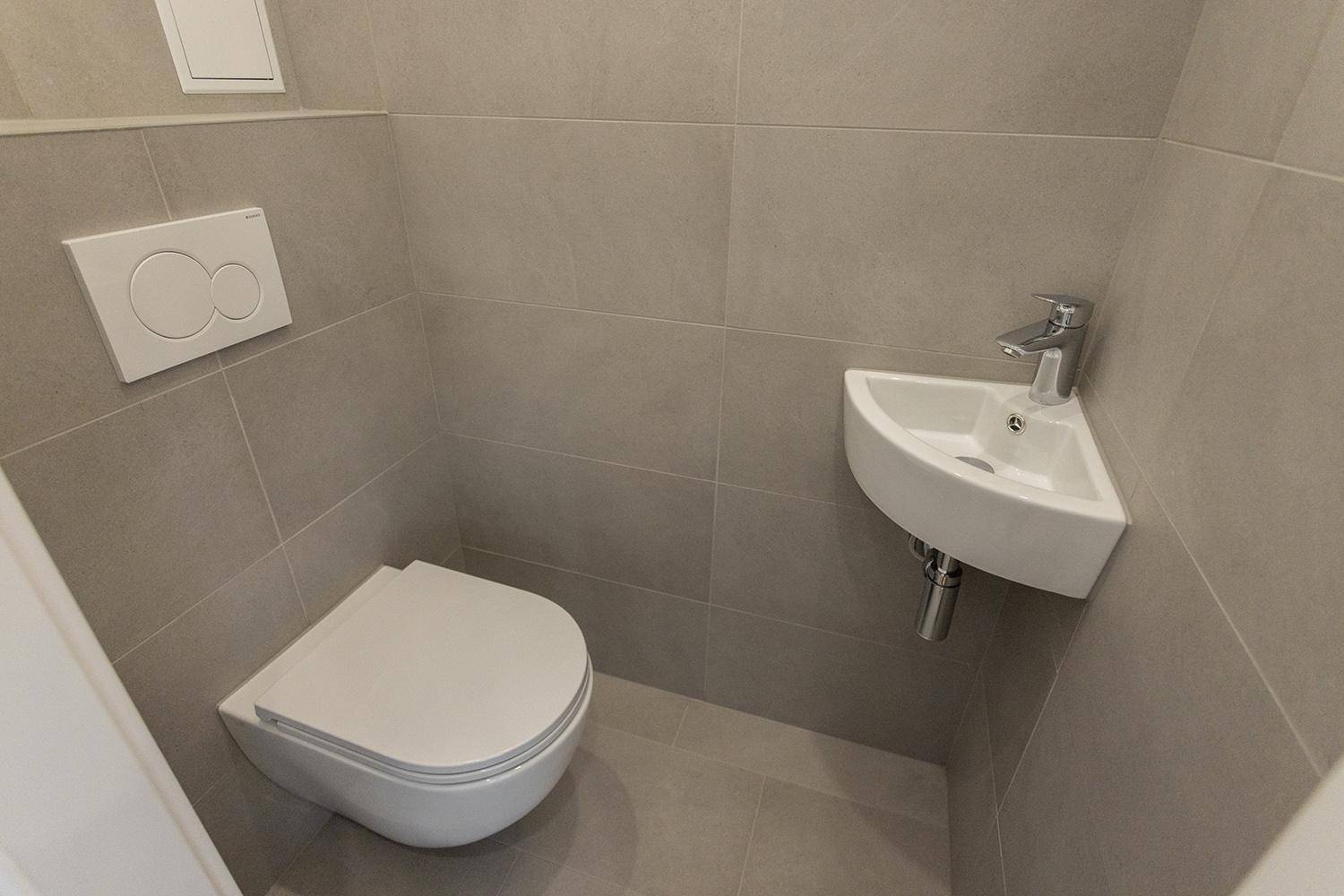 Separates Gäste-WC mit Wand-WC, kleinem Eckwaschbecken und grauen Fliesen.