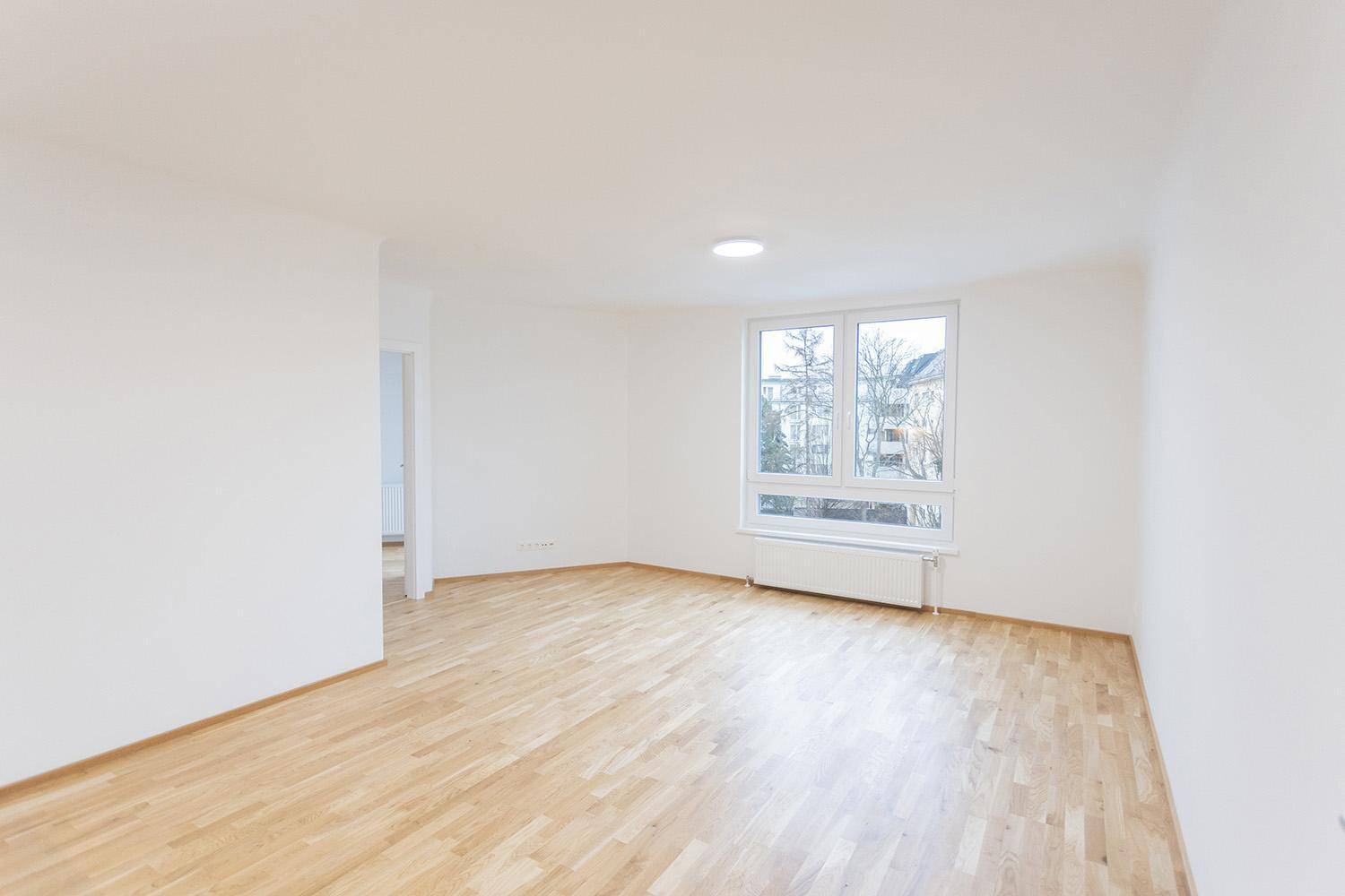 Geräumiges Wohnzimmer mit Holzboden und großem Fenster, das viel Tageslicht hereinlässt.