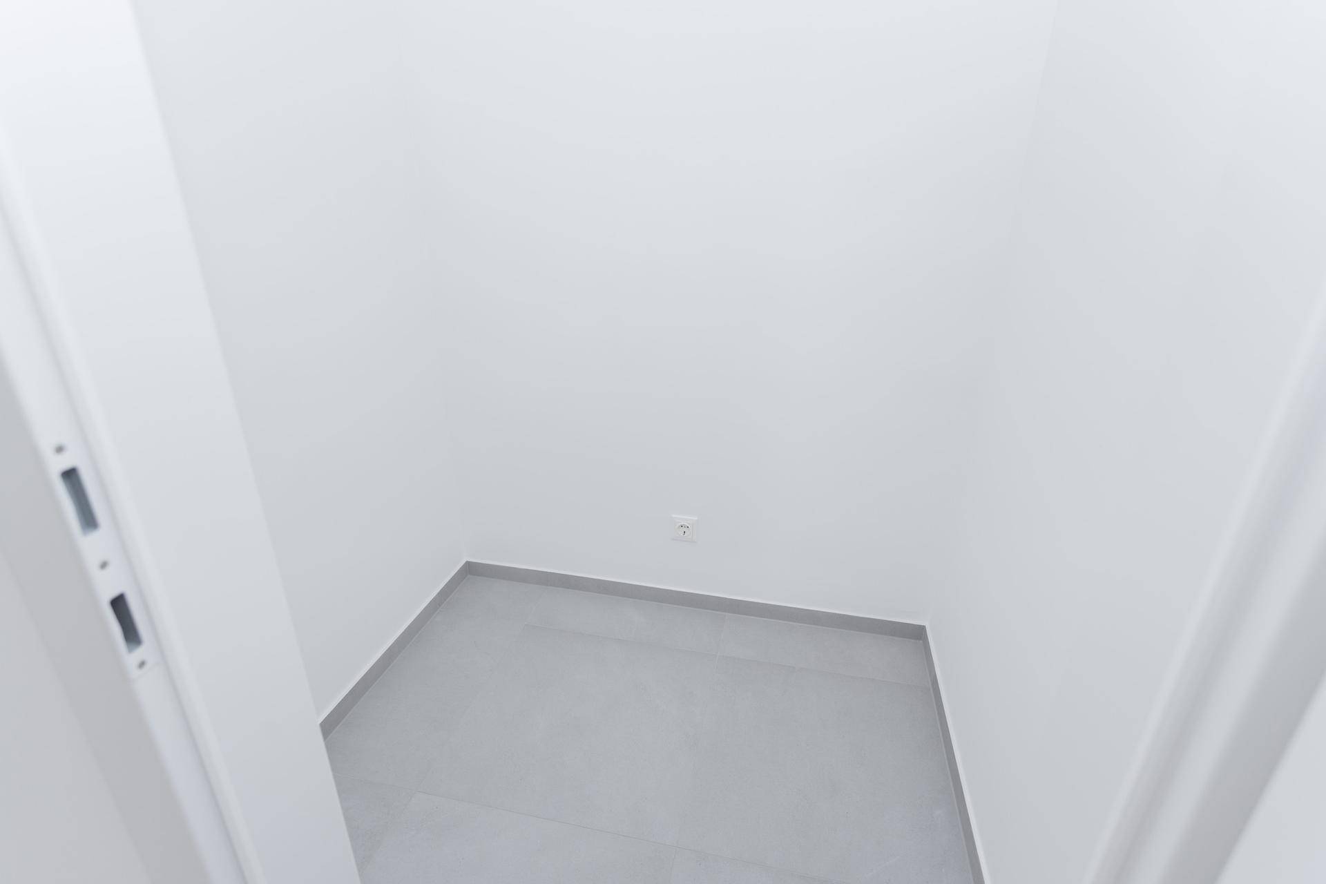 Kleiner, leerer Abstellraum mit grauen Fliesen und einer Steckdose an der Wand.