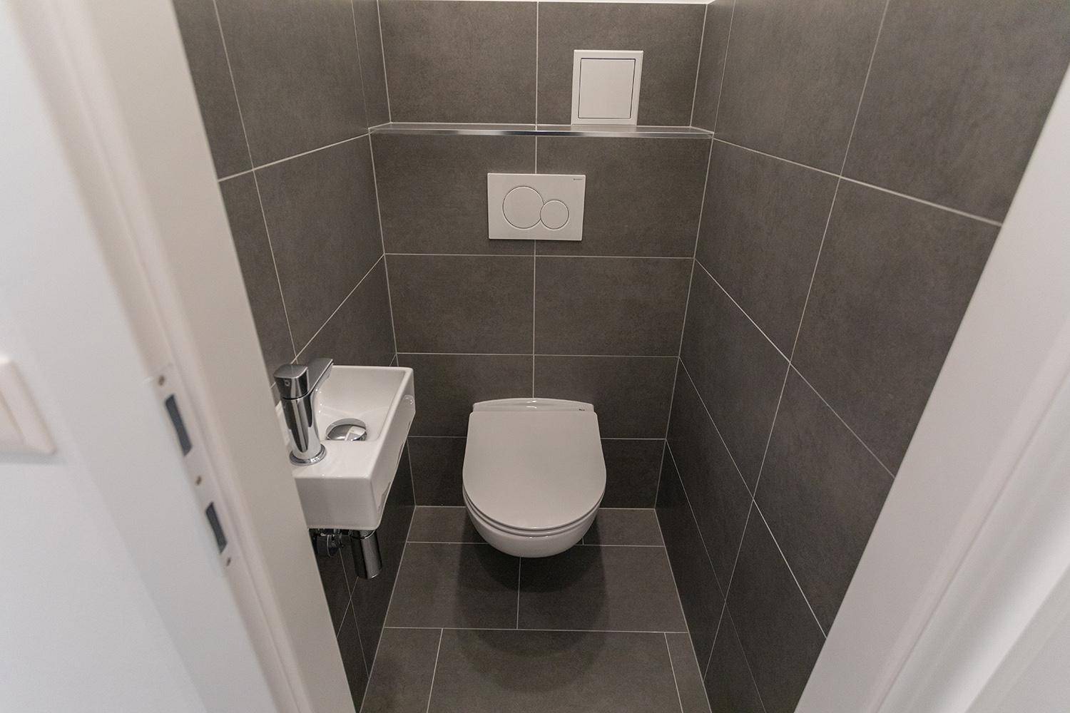 Modernes Gäste-WC mit grauen Fliesen, wandhängendem WC und kleinem Waschbecken.
